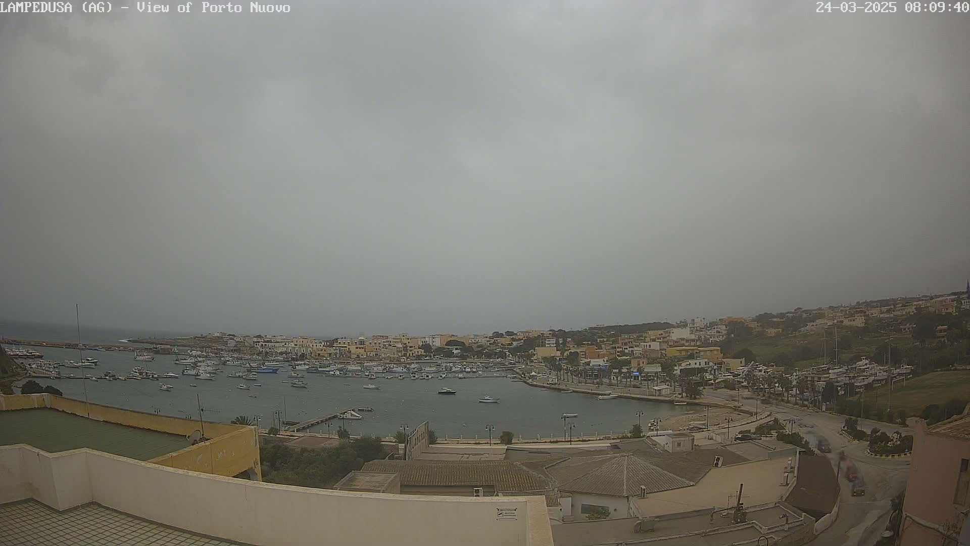 An overcast day reveals a harbor filled with numerous boats, surrounded by a town with low-rise buildings under a gray sky.