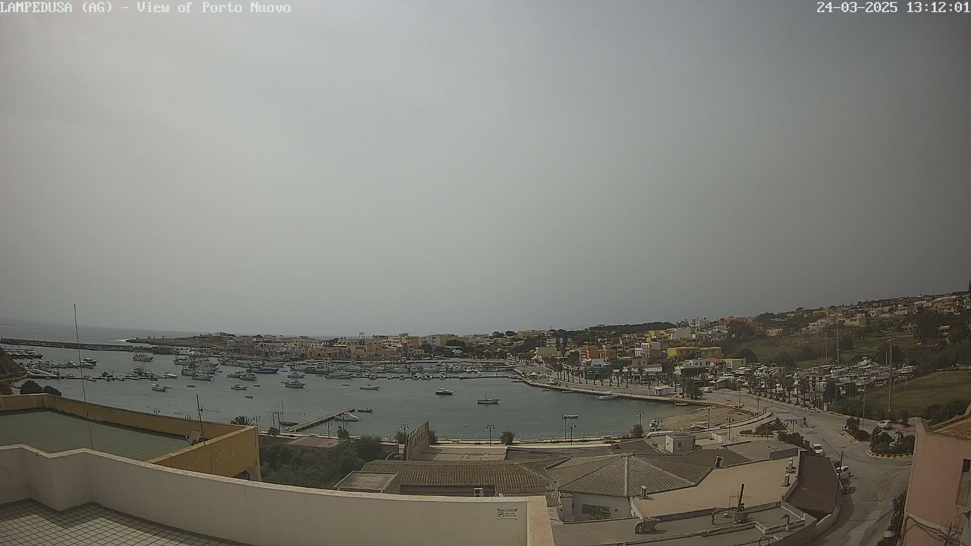 A hazy, overcast day reveals a harbor filled with numerous boats, surrounded by a town with low-rise buildings extending up a hillside.