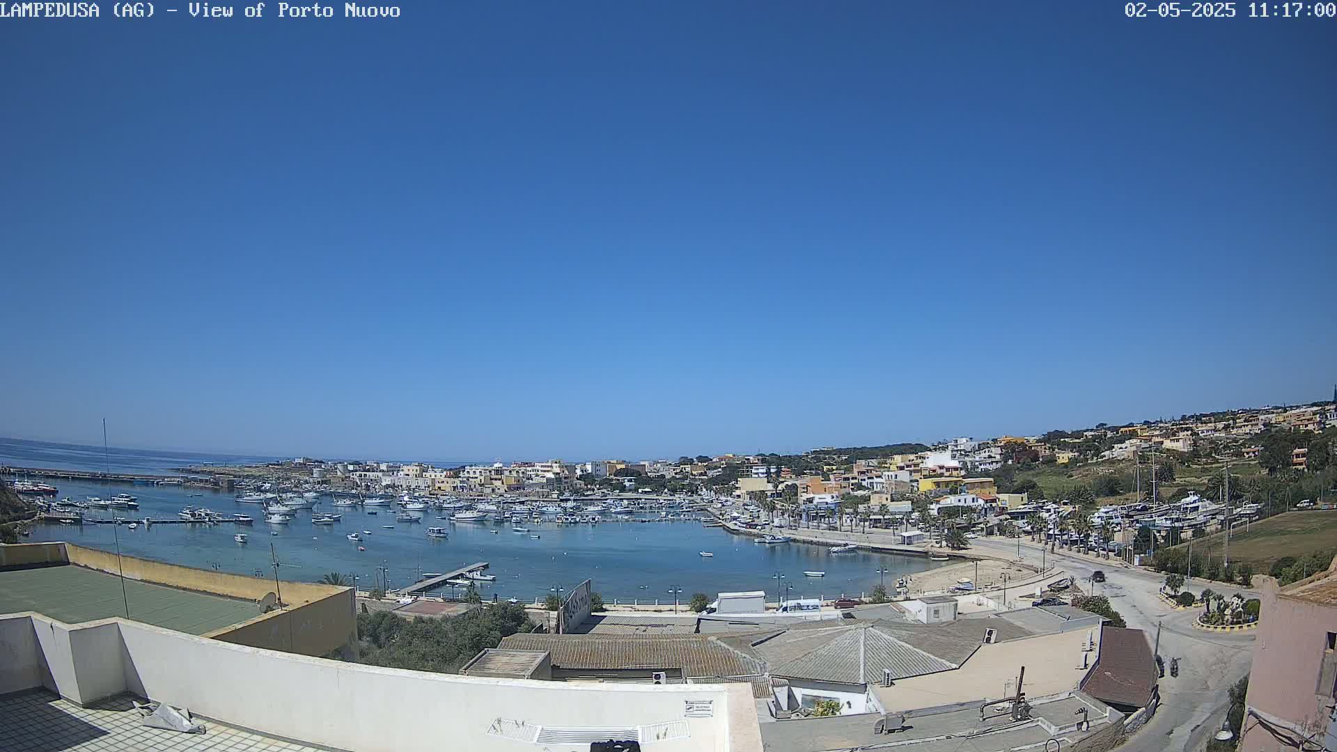 A clear, sunny day overlooks a harbor filled with boats, surrounded by a town of low-rise buildings nestled against a hillside.