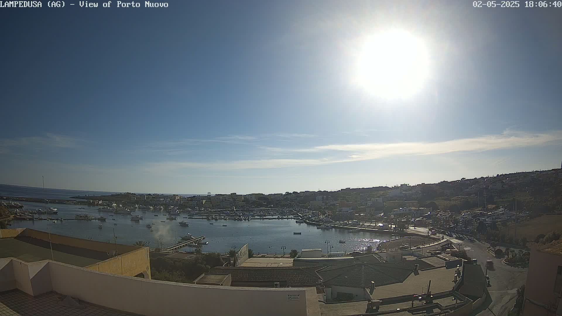 A sunny harbor scene shows a calm body of water filled with boats, surrounded by buildings and hills under a mostly clear sky.