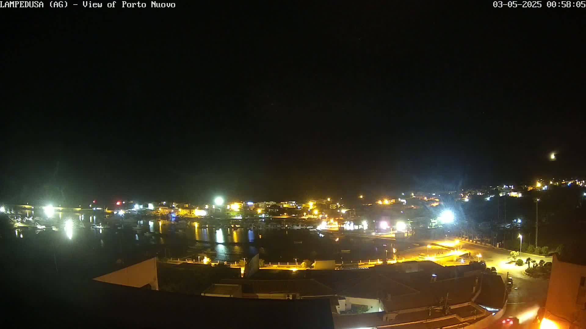 A nighttime, overhead view of a harbor town with many lights illuminating boats and buildings under a clear, dark sky.