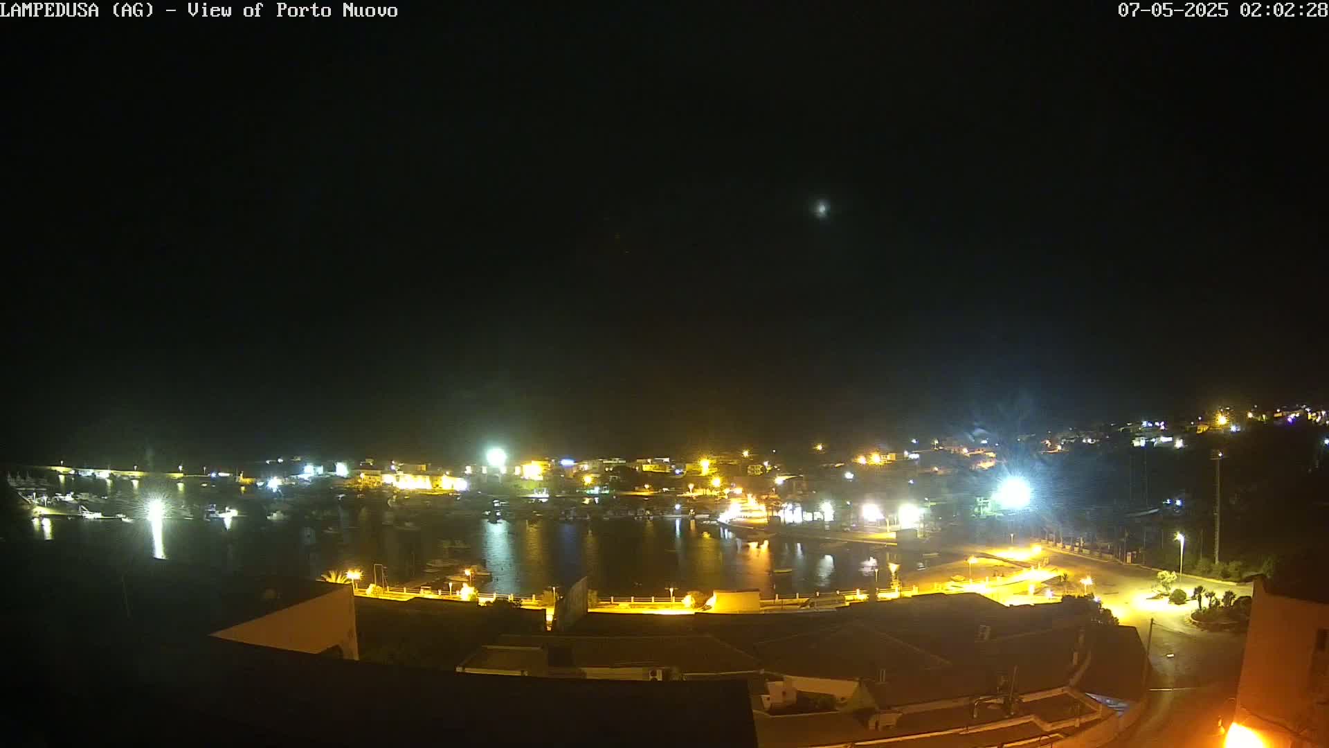 A nighttime, aerial view of a harbor town with boats in the water, illuminated by many lights under a clear, dark sky.