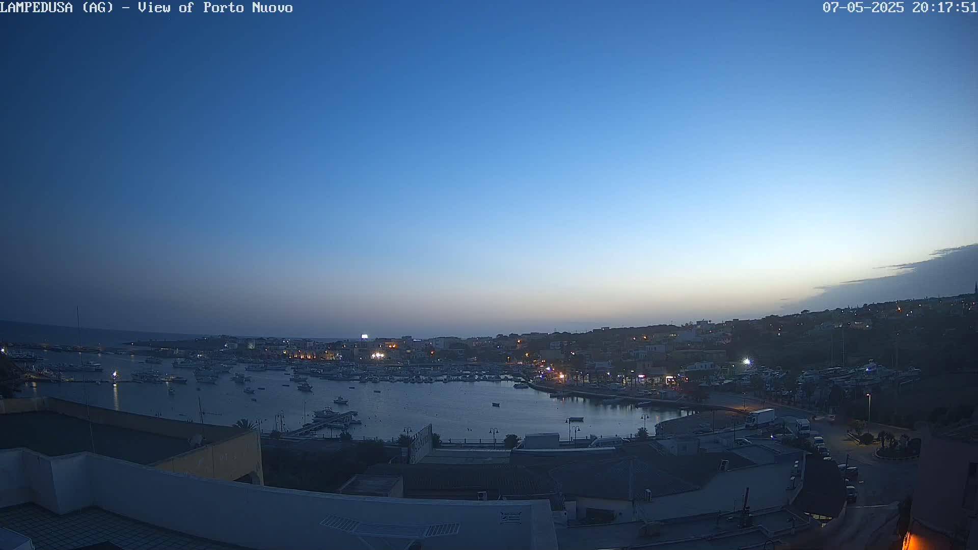 A harbor filled with boats is nestled in a town at twilight under a clear blue sky.