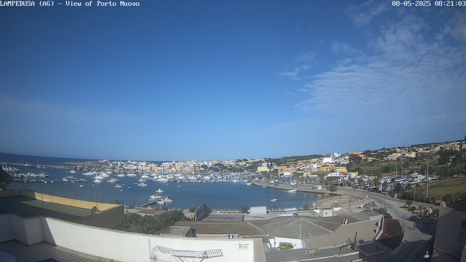 A mostly sunny day overlooks a harbor filled with boats, adjacent to a town of light-colored buildings on a hillside.