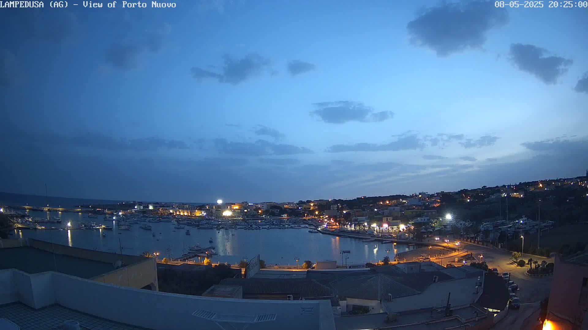 A nighttime view of a harbor filled with boats, surrounded by a town with lights illuminated against a twilight sky with a few clouds.