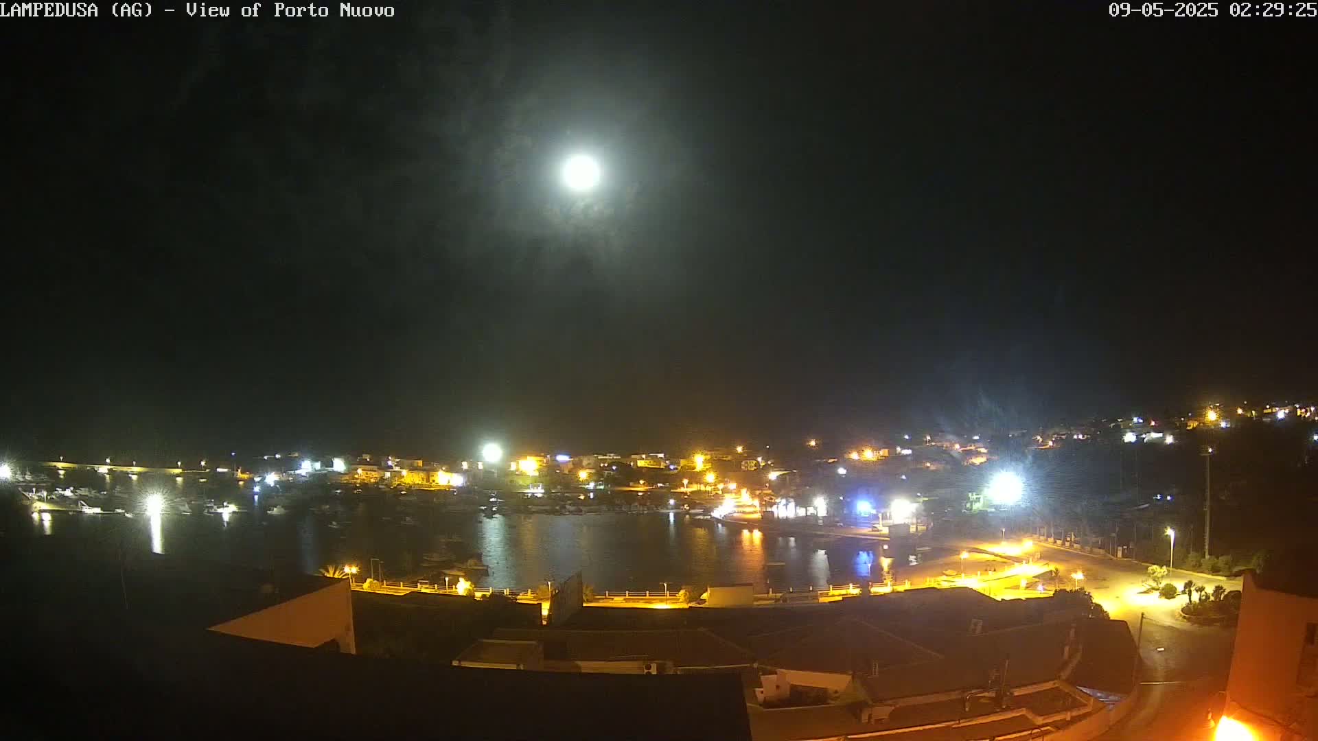A brightly lit harbor town is nestled below a night sky with a full moon partially obscured by thin clouds.