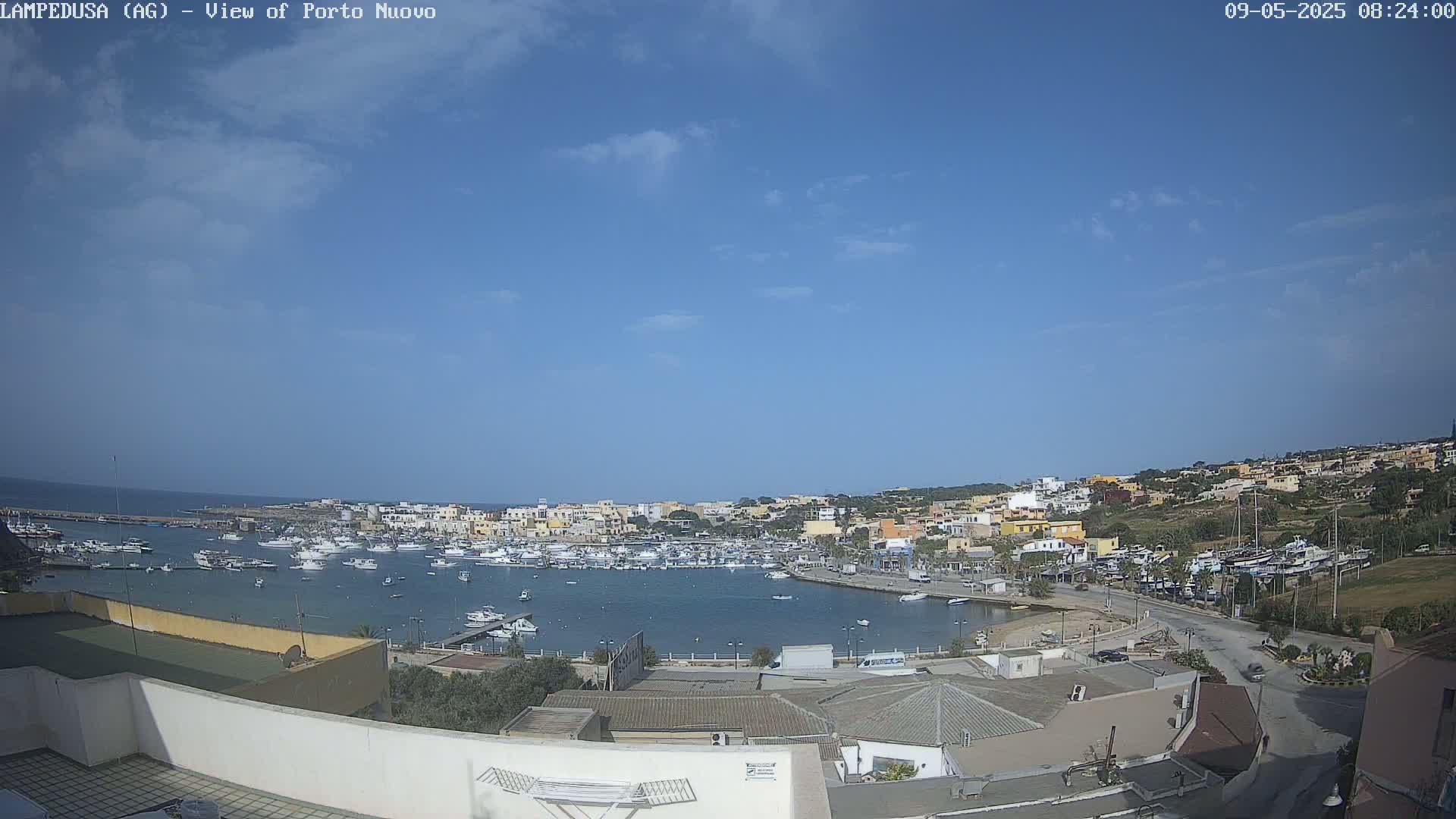 A harbor filled with numerous boats is situated beside a town of low-rise buildings under a mostly clear, bright blue sky.