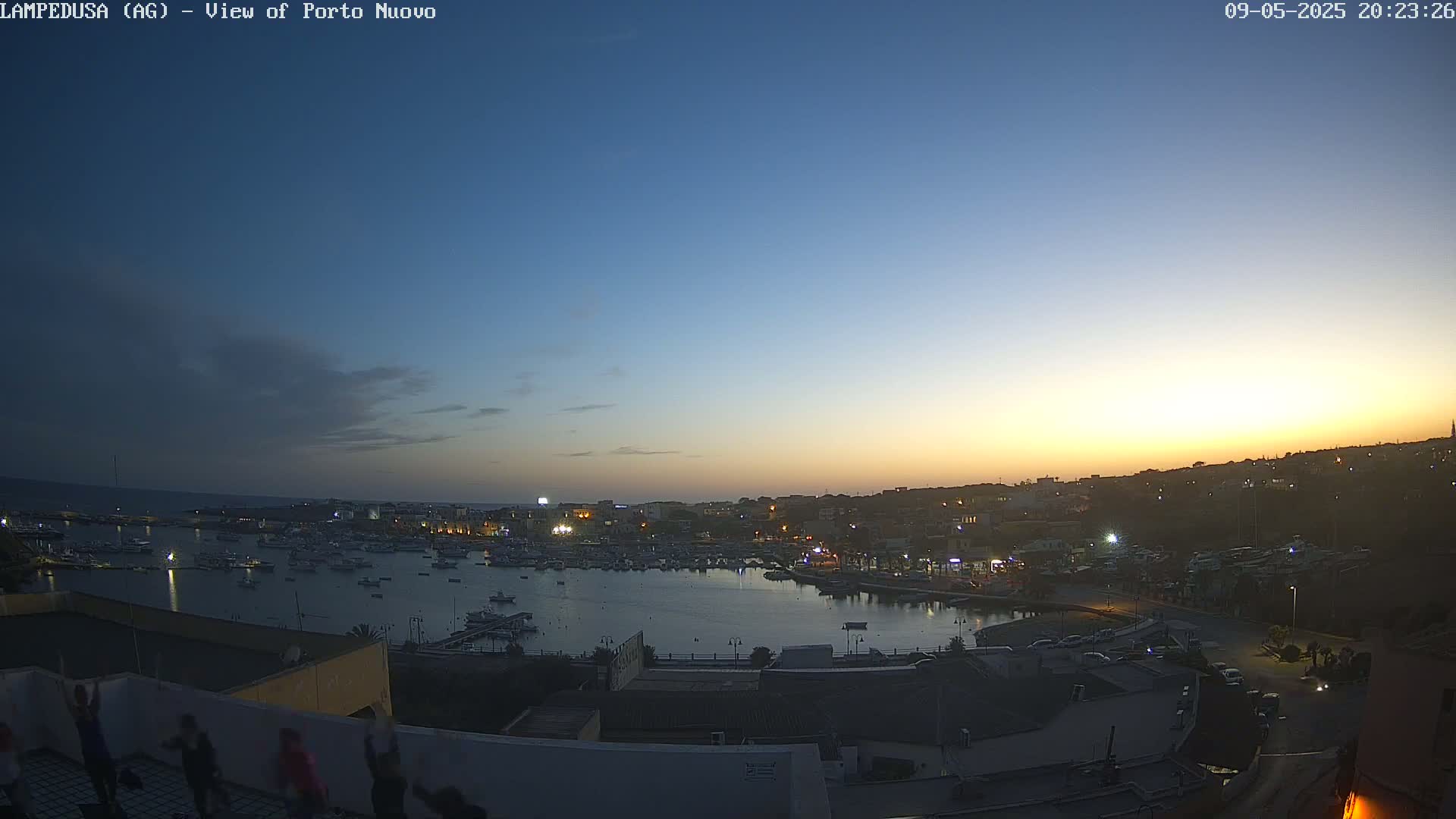 A twilight view of a harbor filled with boats, overlooked by a town illuminated against a clear, dusky sky.