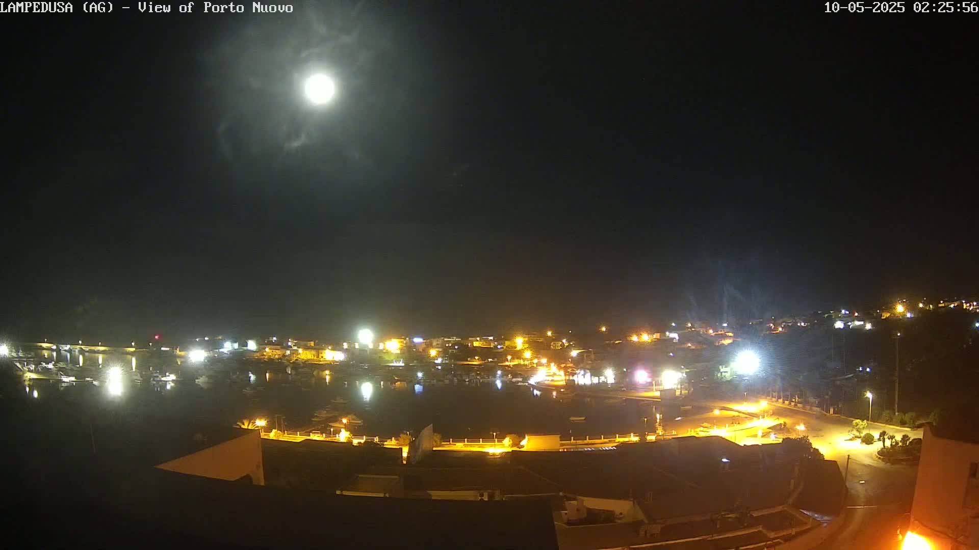 A brightly lit harbor town is visible under a nighttime sky with a full moon partially obscured by clouds.