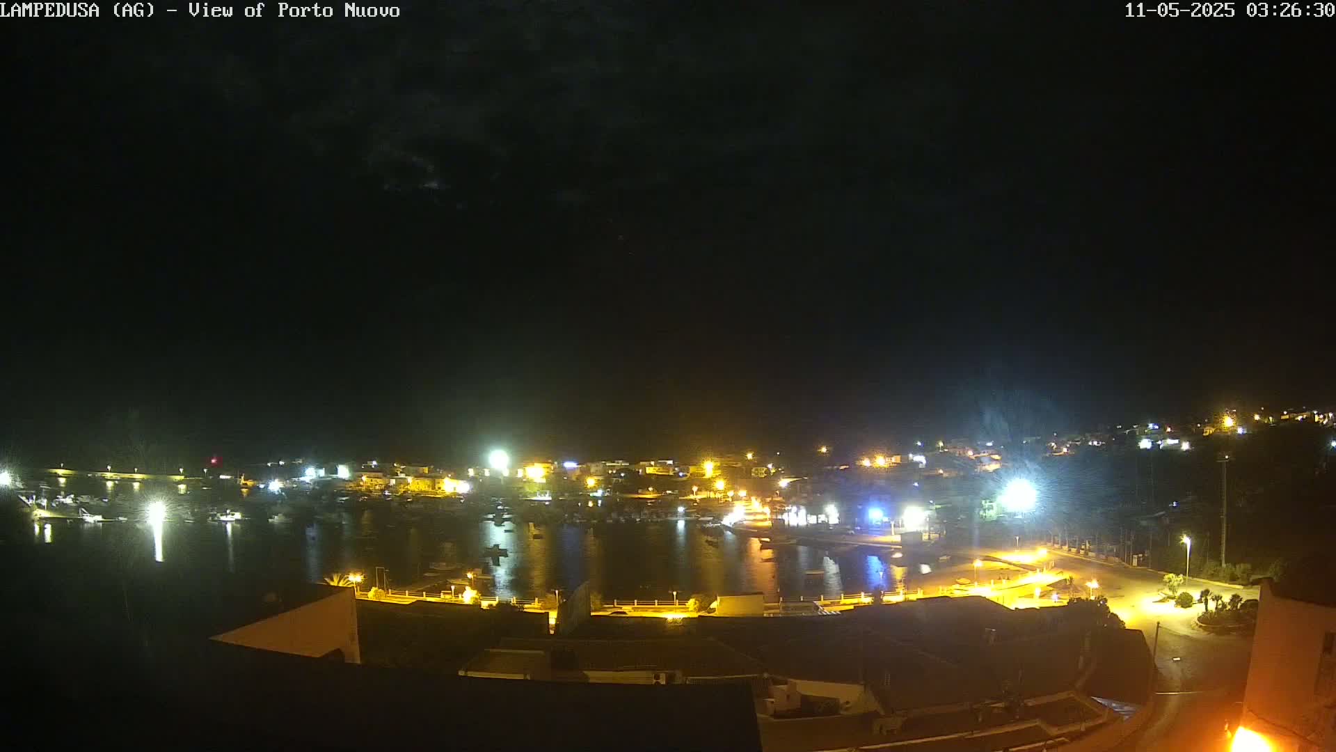 A nighttime view of a harbor town with its lights illuminating the water and buildings under a mostly clear sky.