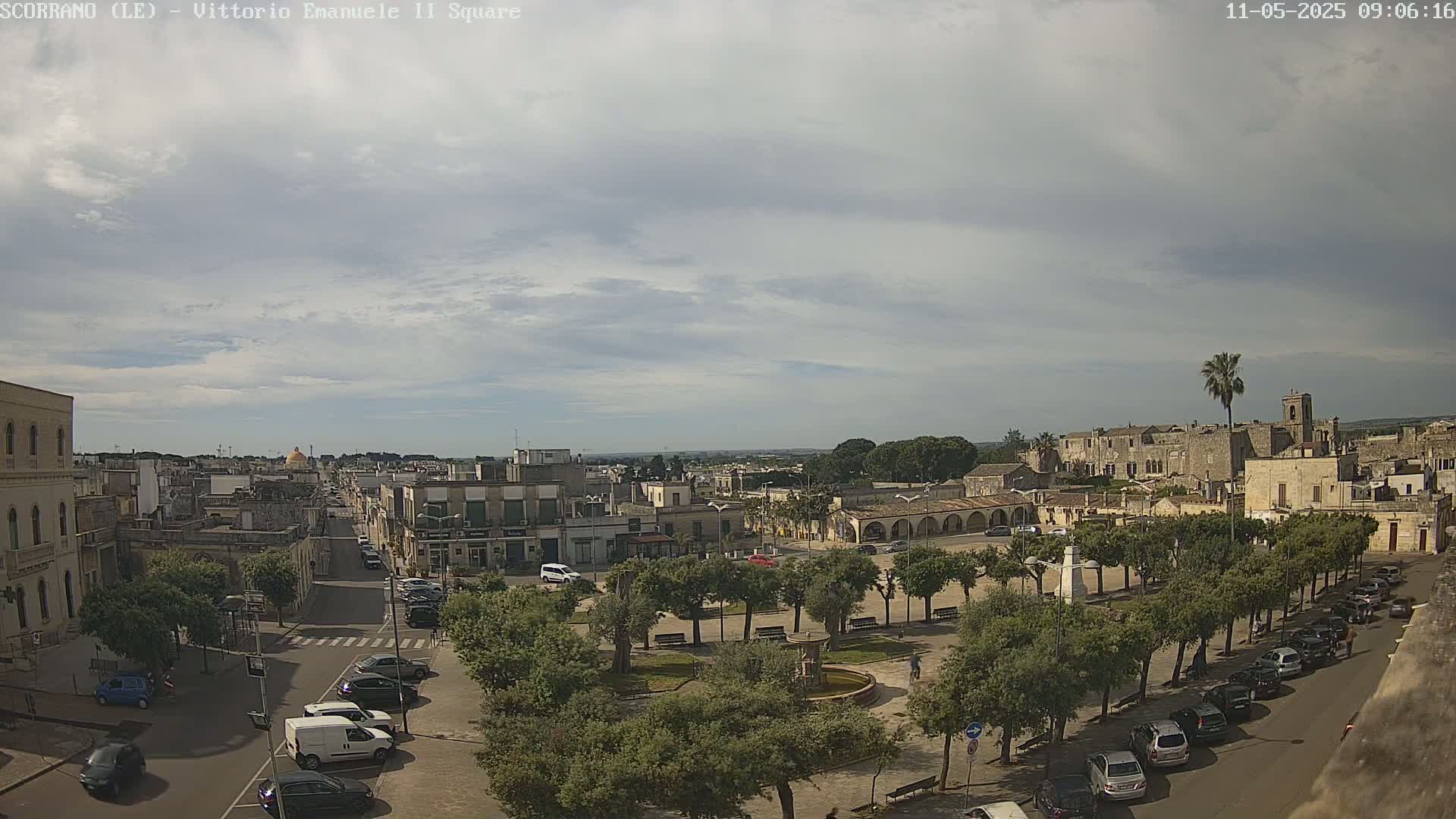 A mostly cloudy day overlooks a town square with a fountain, lined with trees and surrounded by buildings and parked cars.