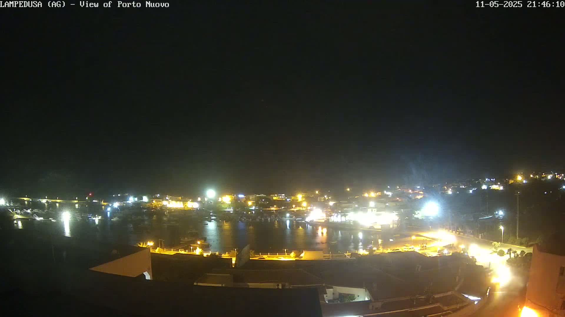 A nighttime, aerial view shows a harbor filled with boats, surrounded by a brightly lit town under a clear, dark sky.