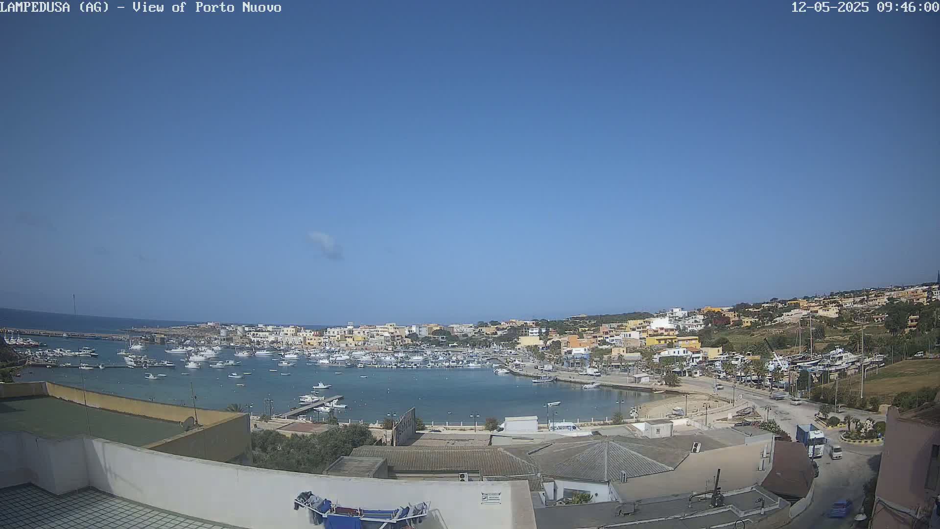 A harbor filled with numerous boats is nestled within a town of low-rise buildings under a clear blue sky.