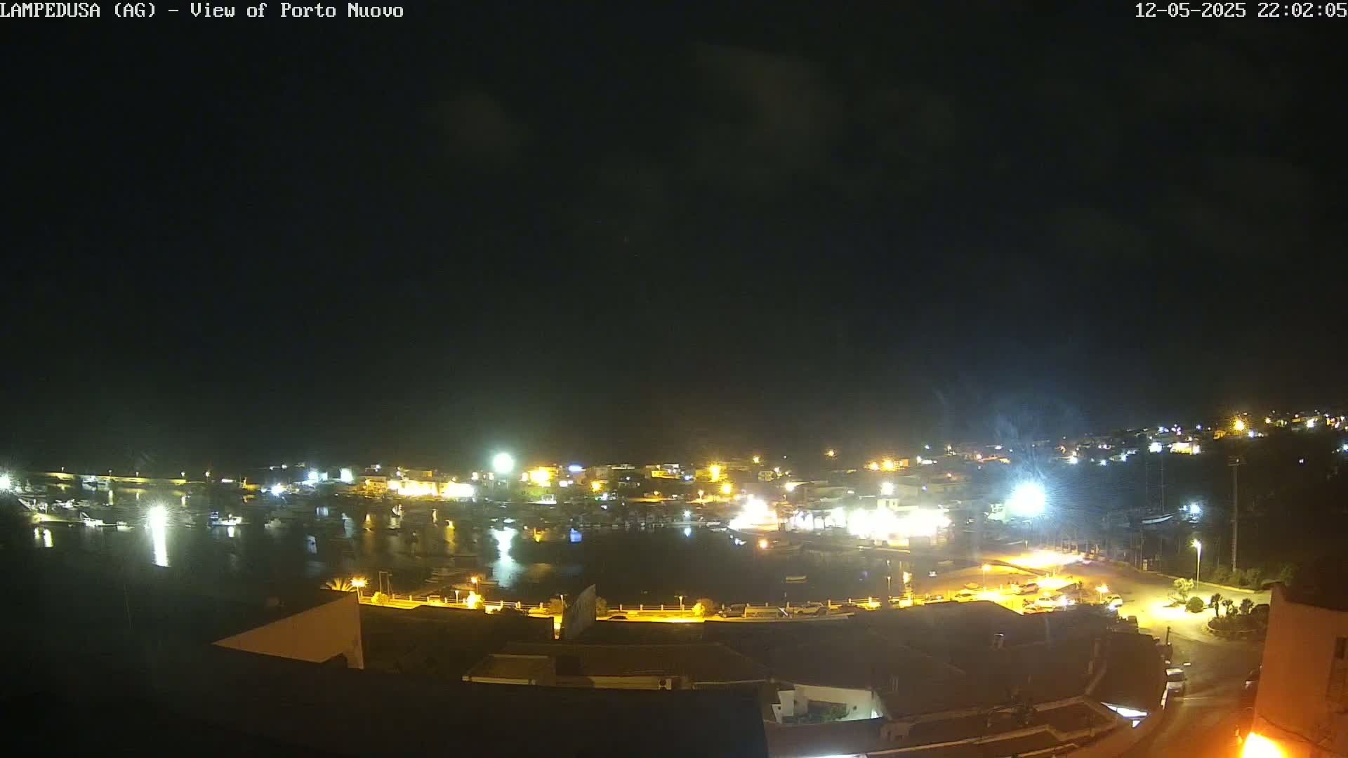 A nighttime view of a harbor town with many lights illuminating the buildings and boats, under a mostly clear sky.