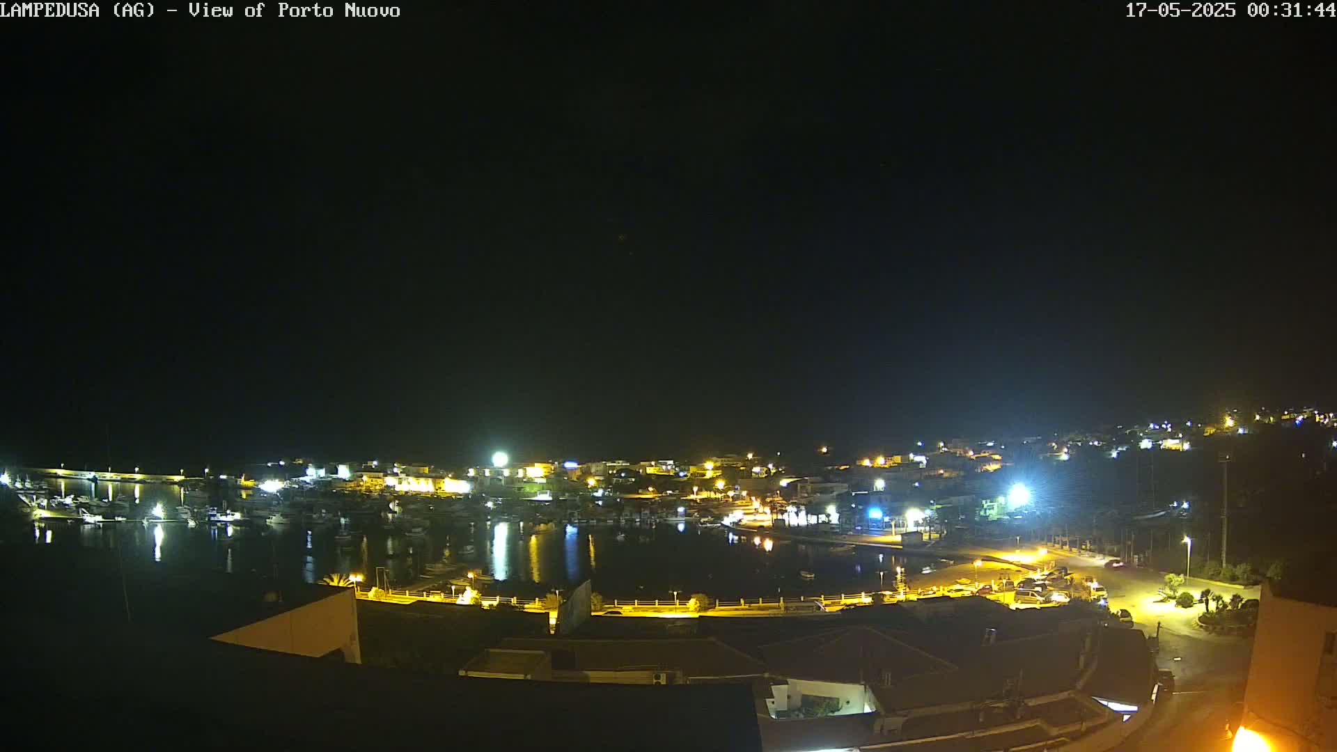 A nighttime view of a harbor town, with boats in the water and buildings illuminated along the shoreline under a clear, dark sky.
