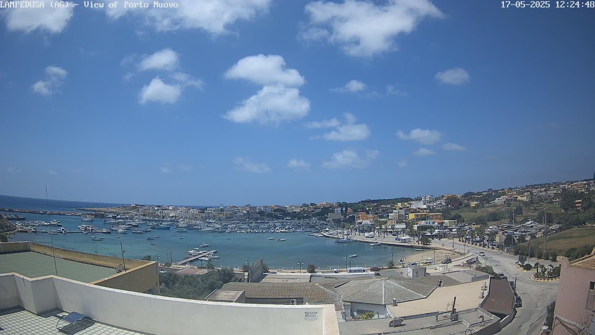 A harbor filled with boats is nestled within a town on a sunny day with a few scattered clouds.