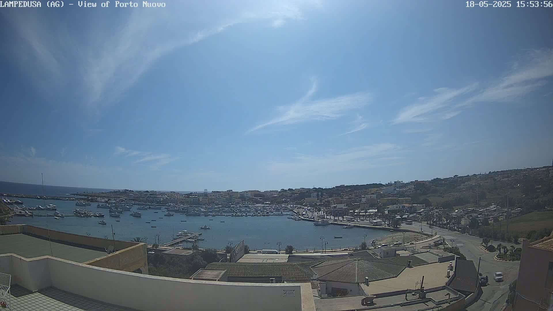 An elevated view of a harbor filled with boats, under a mostly sunny sky with a few scattered clouds, shows a town built on a hillside overlooking the water.