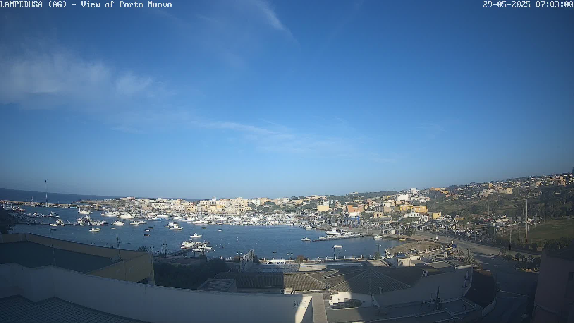 A mostly sunny day reveals a harbor filled with numerous boats, nestled within a town of low-lying buildings that extend up a hillside.