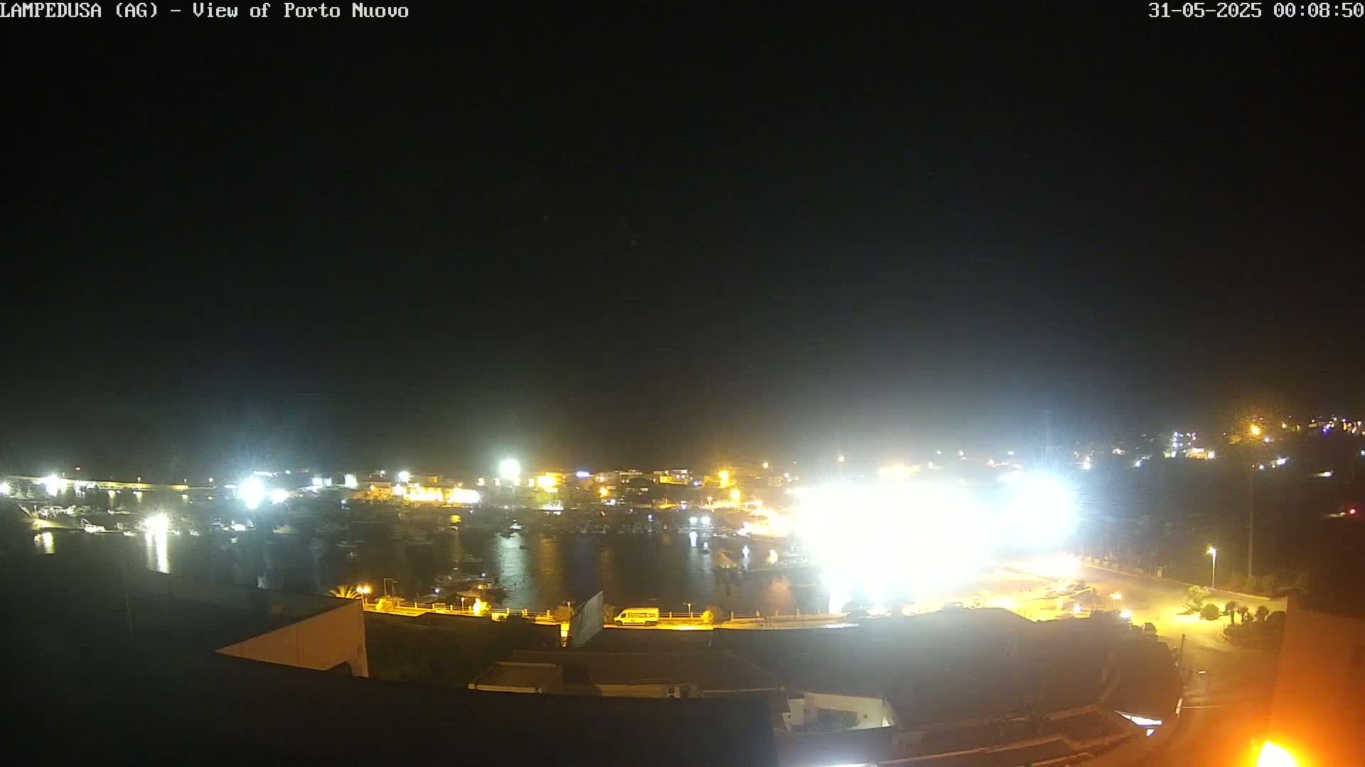 A brightly lit harbor at night is shown, with boats moored in the water and buildings surrounding it.