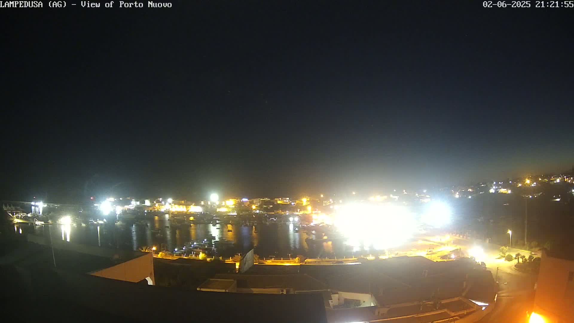 A brightly lit harbor at night, filled with boats, is viewed from a slightly elevated vantage point under a clear, dark sky.