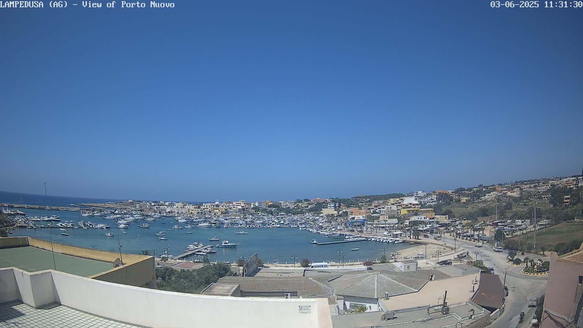 A clear, sunny day overlooks a harbor filled with numerous boats, surrounded by a town of low-rise buildings nestled against a hillside.