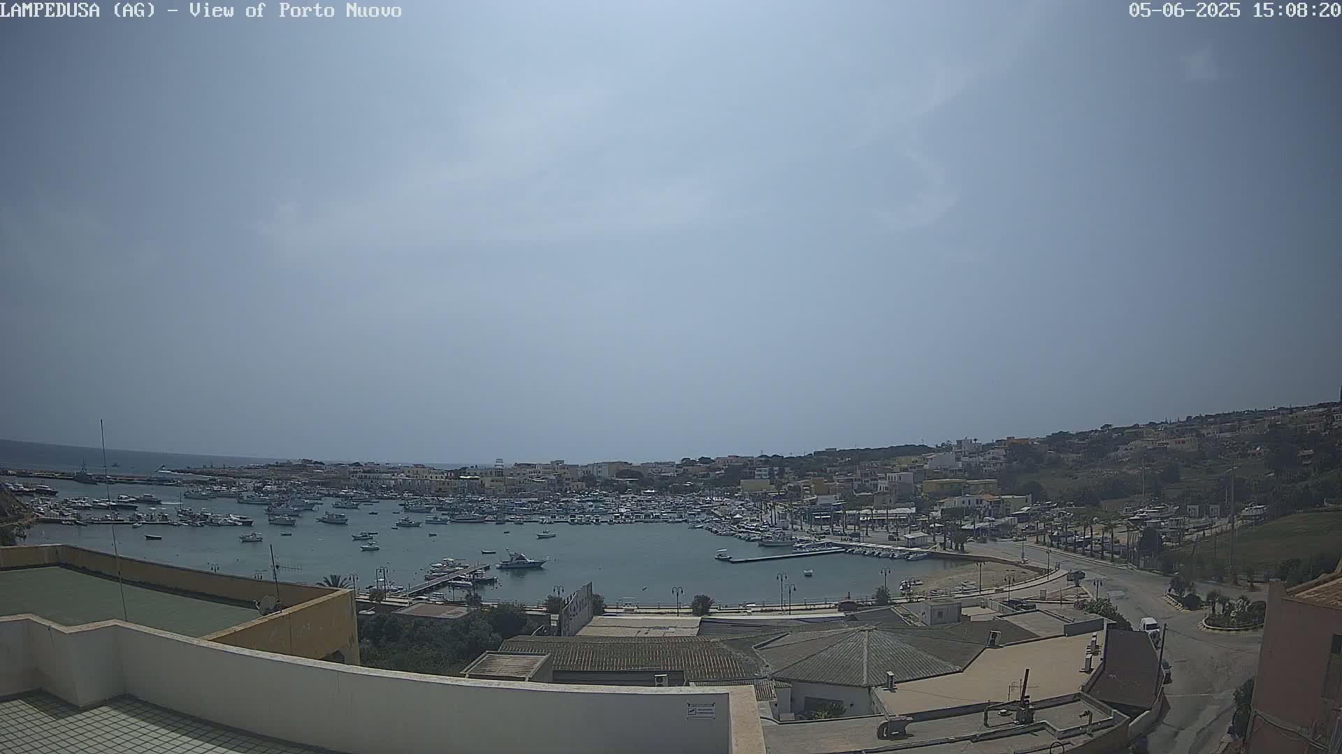 A mostly sunny day shows a harbor filled with numerous boats, adjacent to a town built on a hillside overlooking the water.
