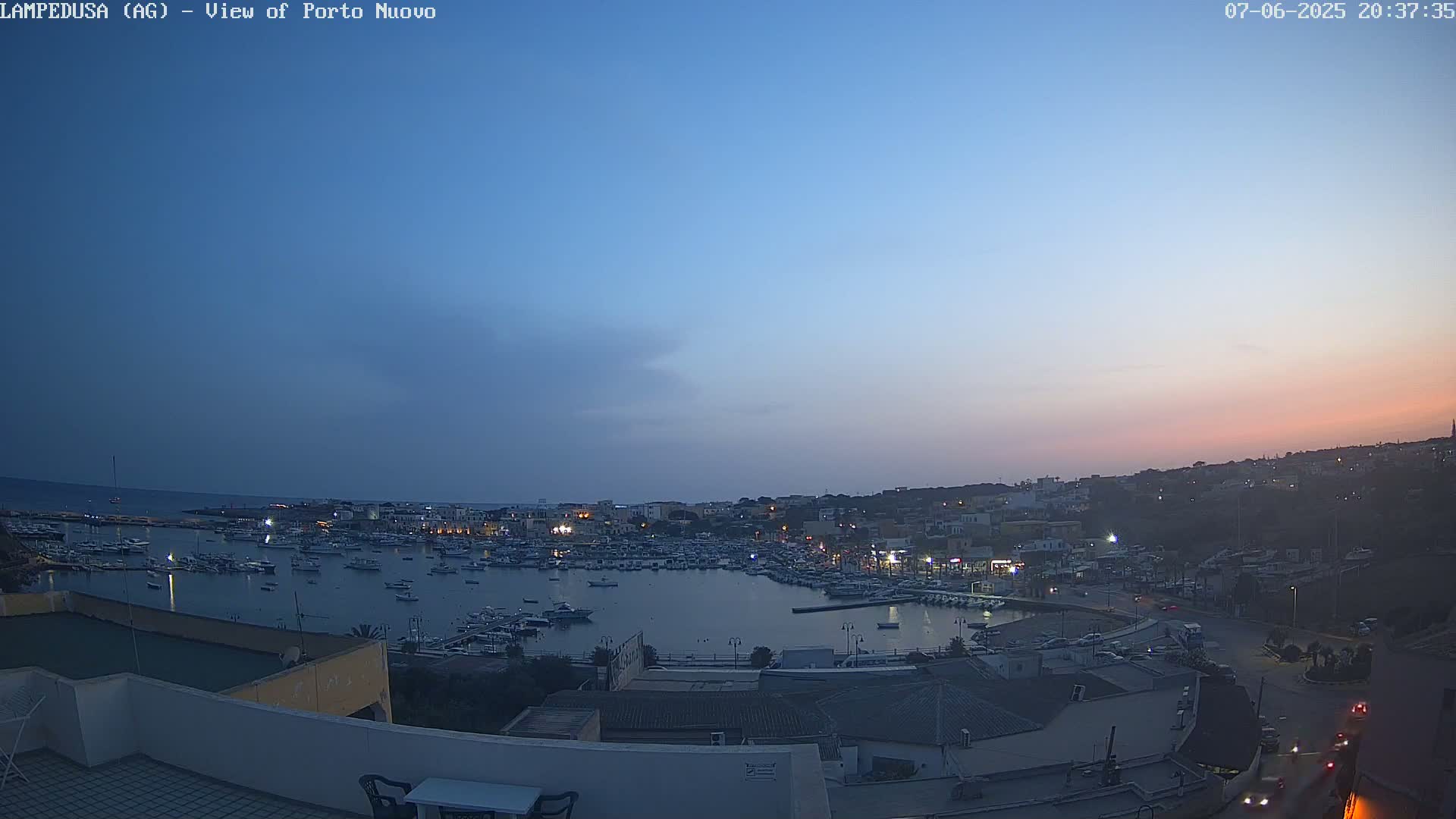 An evening harbor scene shows a calm body of water filled with boats, surrounded by a town with buildings lit up against a twilight sky.
