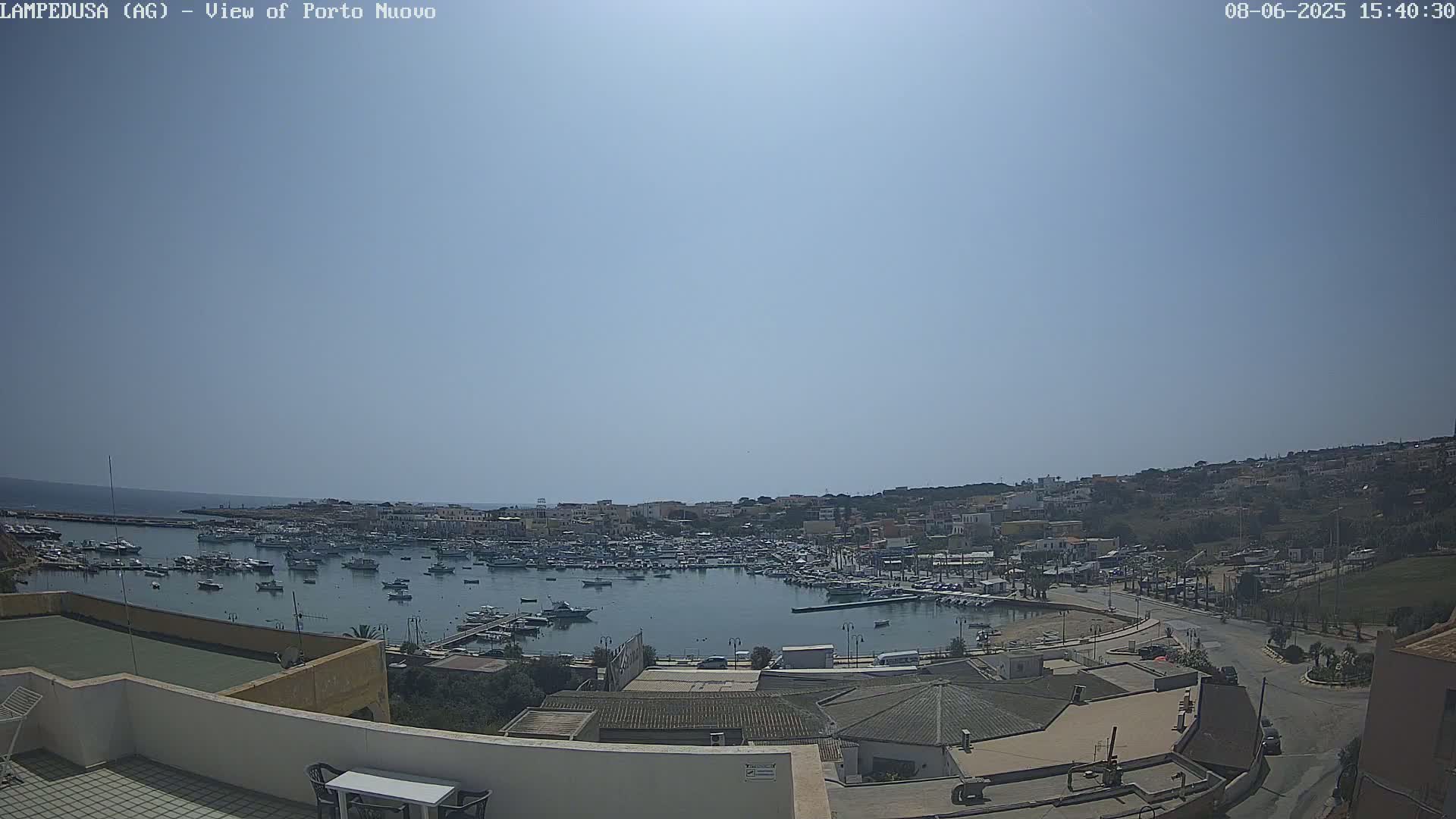 A sunny day overlooking a harbor filled with numerous boats, surrounded by a town built on a hillside, extending to a calm sea in the background.