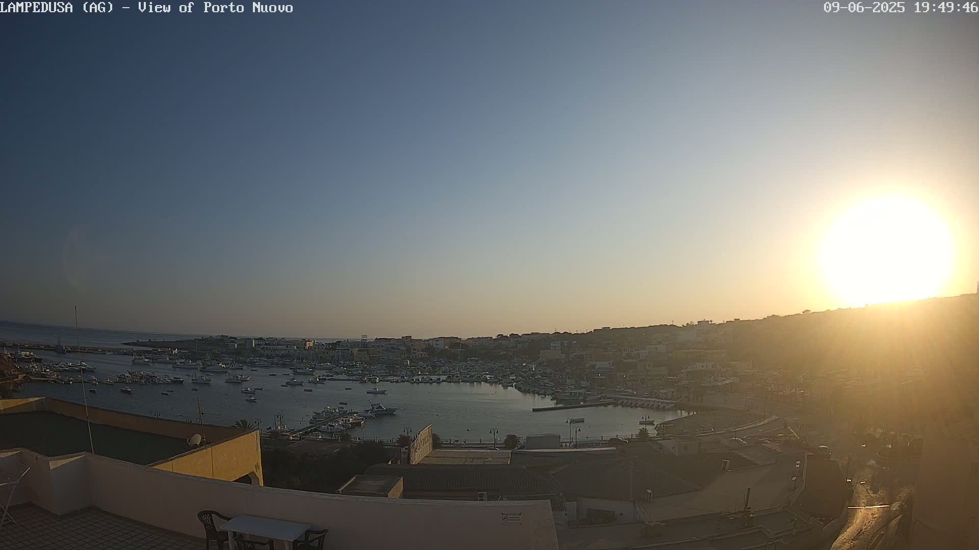 A sun-drenched harbor filled with boats is viewed from an elevated position, with the bright sun partially obscured by atmospheric haze.