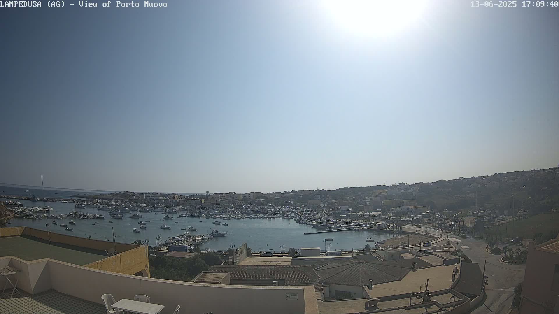 A sunny day overlooking a harbor filled with boats, a town nestled on a hillside, and a rooftop patio with outdoor furniture.