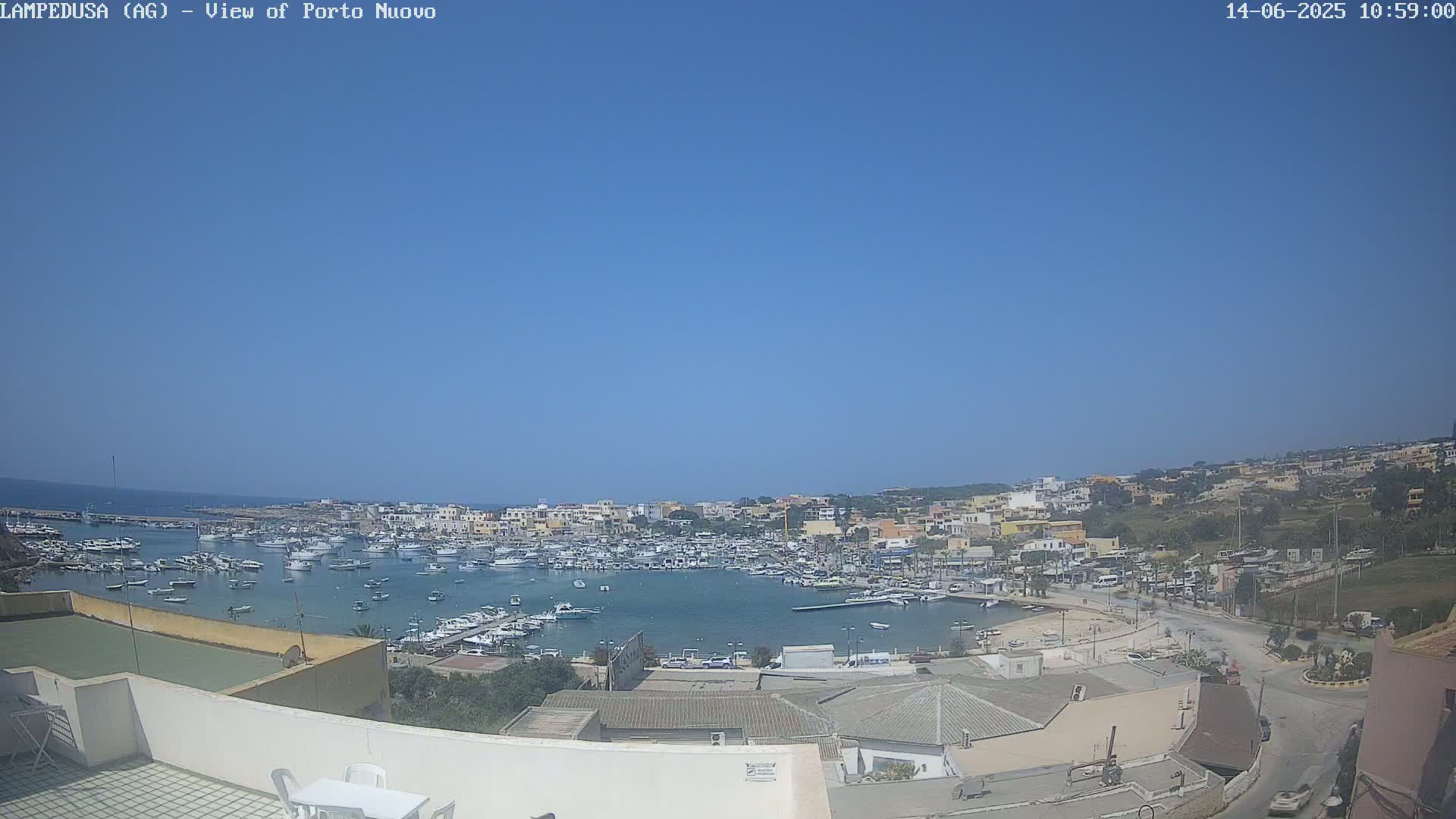A harbor filled with numerous boats is visible under a clear blue sky, with a town situated on a hillside overlooking the water.