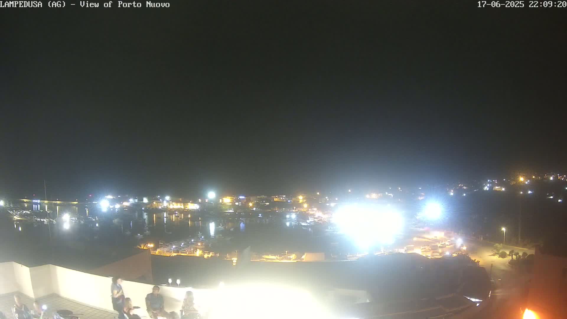 A nighttime view from an elevated position shows a harbor filled with boats and surrounded by illuminated buildings under a clear, dark sky.