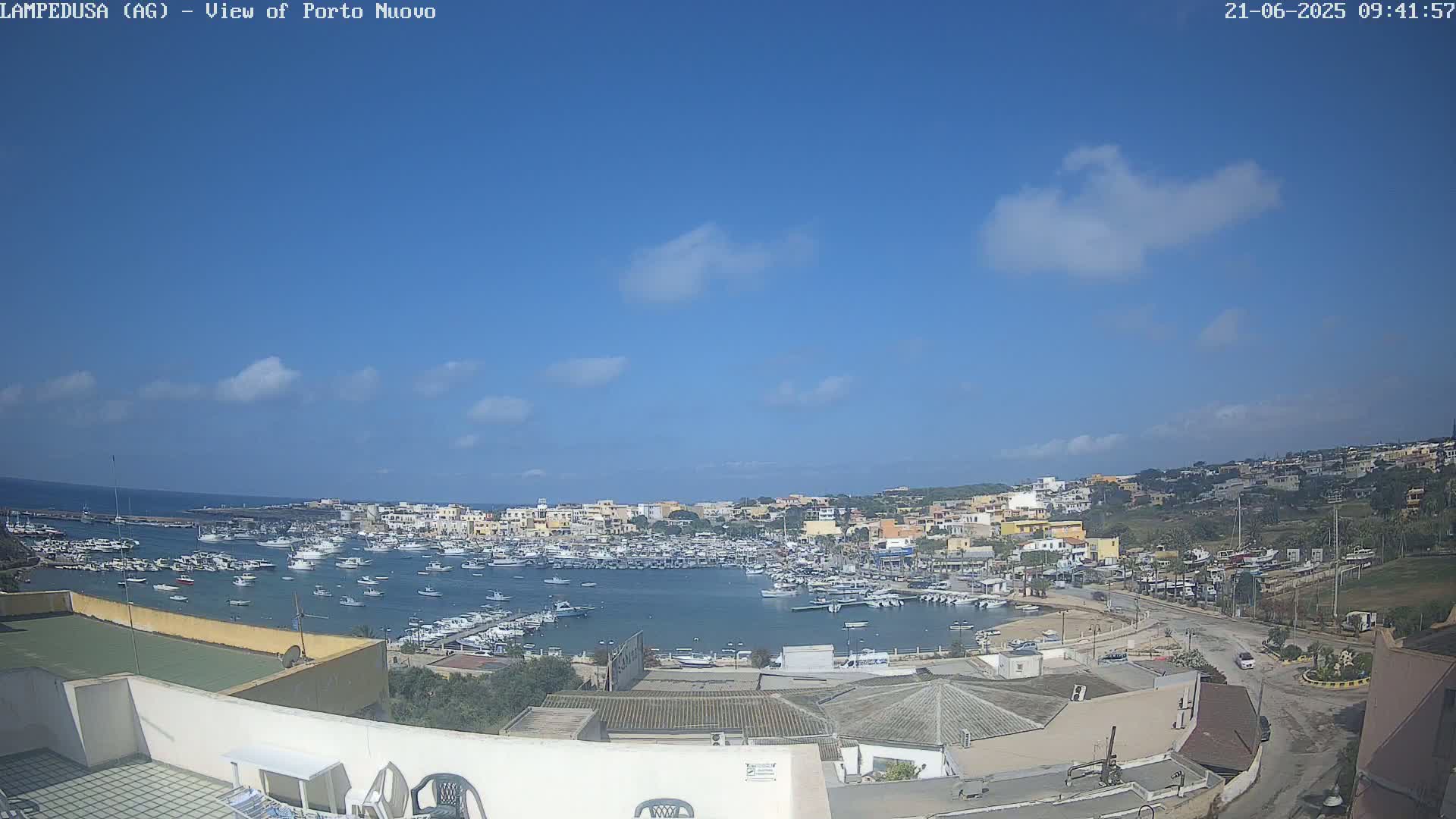 A mostly sunny day overlooks a harbor filled with boats and a town of low-rise buildings nestled along a coastline.
