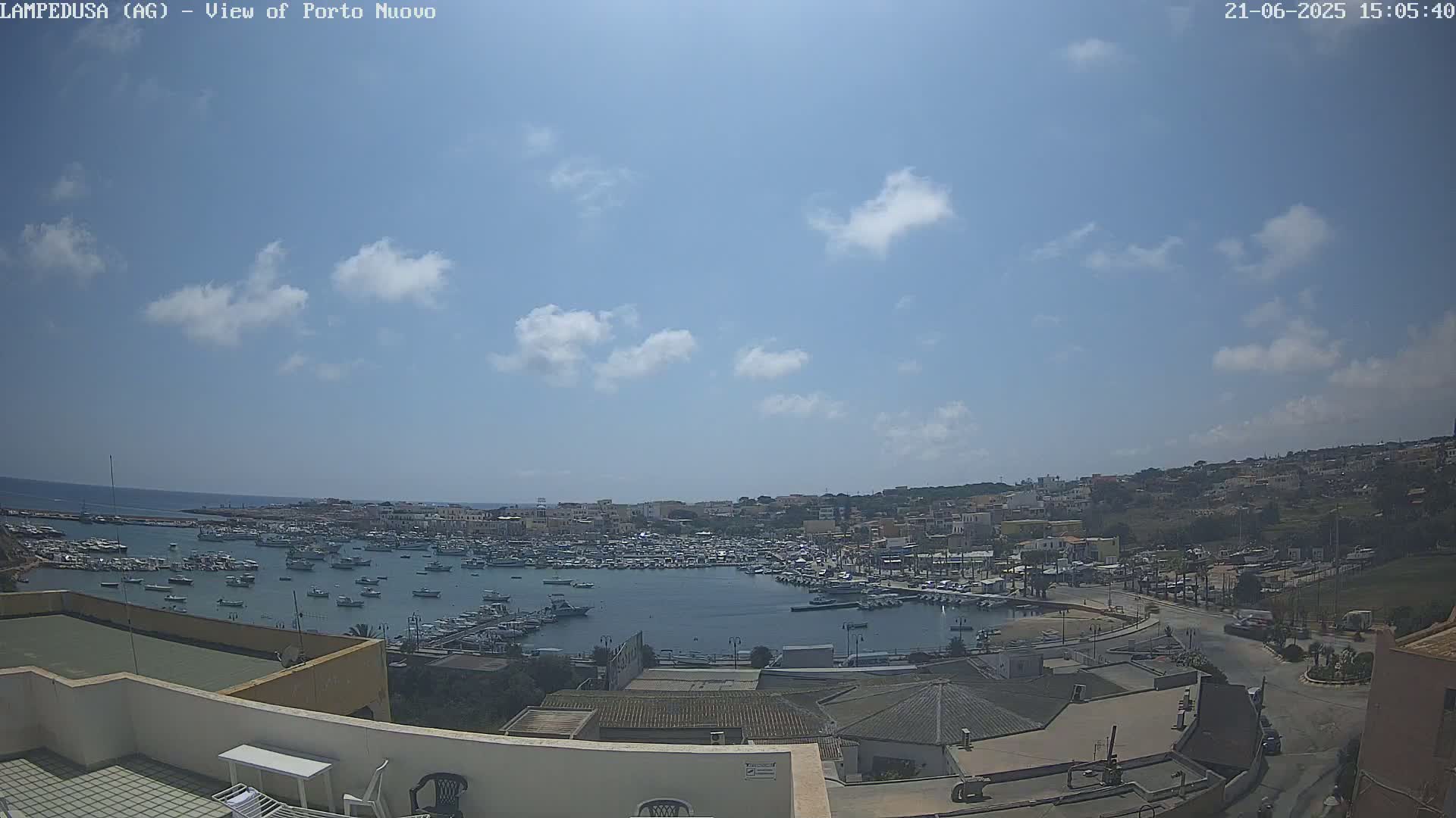 A harbor filled with numerous boats is nestled within a town situated on a hill overlooking the ocean under a mostly sunny sky with scattered clouds.
