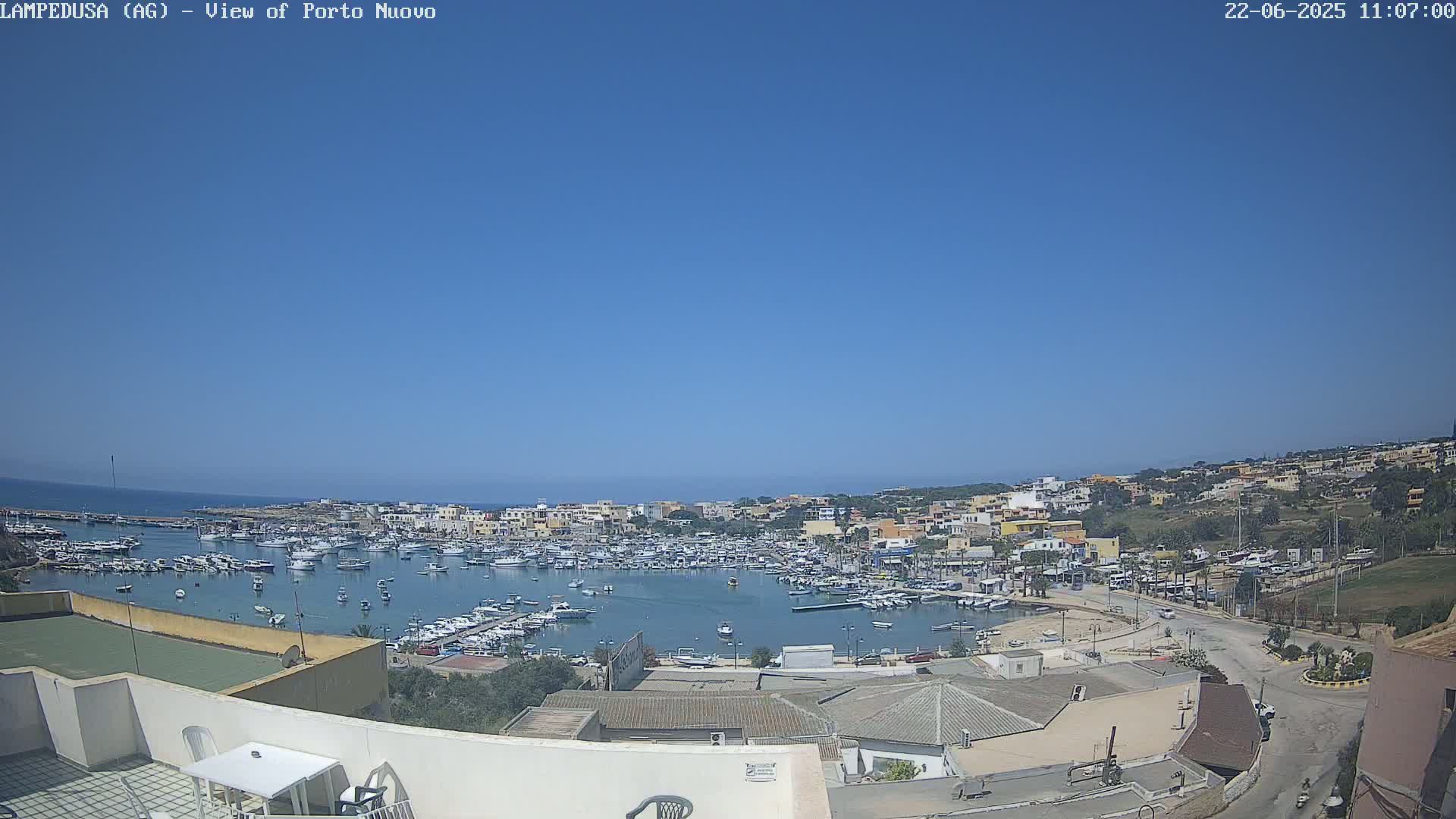 A harbor filled with boats is nestled within a town situated on a hill overlooking the ocean under a clear blue sky.