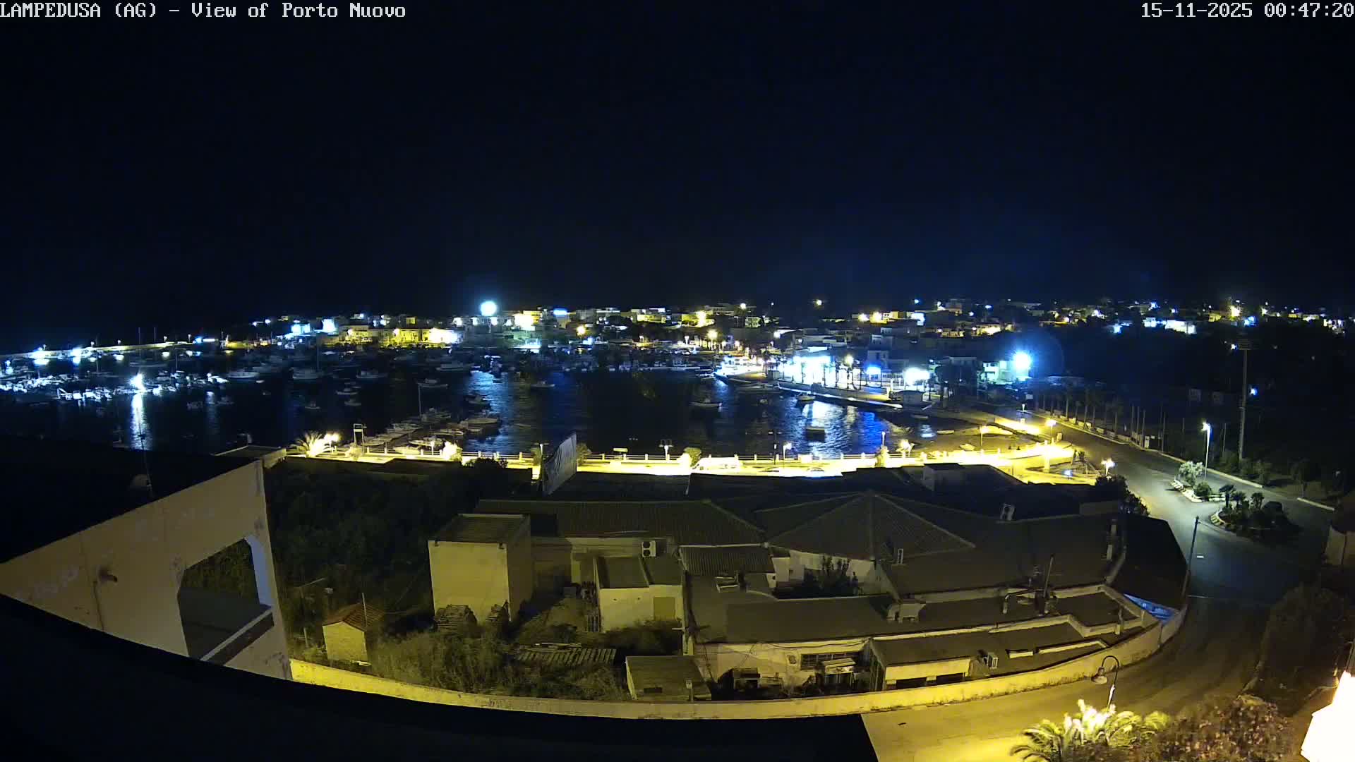 A clear night view shows a brightly illuminated harbor filled with numerous boats docked in calm water, bordered by a town with glowing buildings and streets under a dark sky.