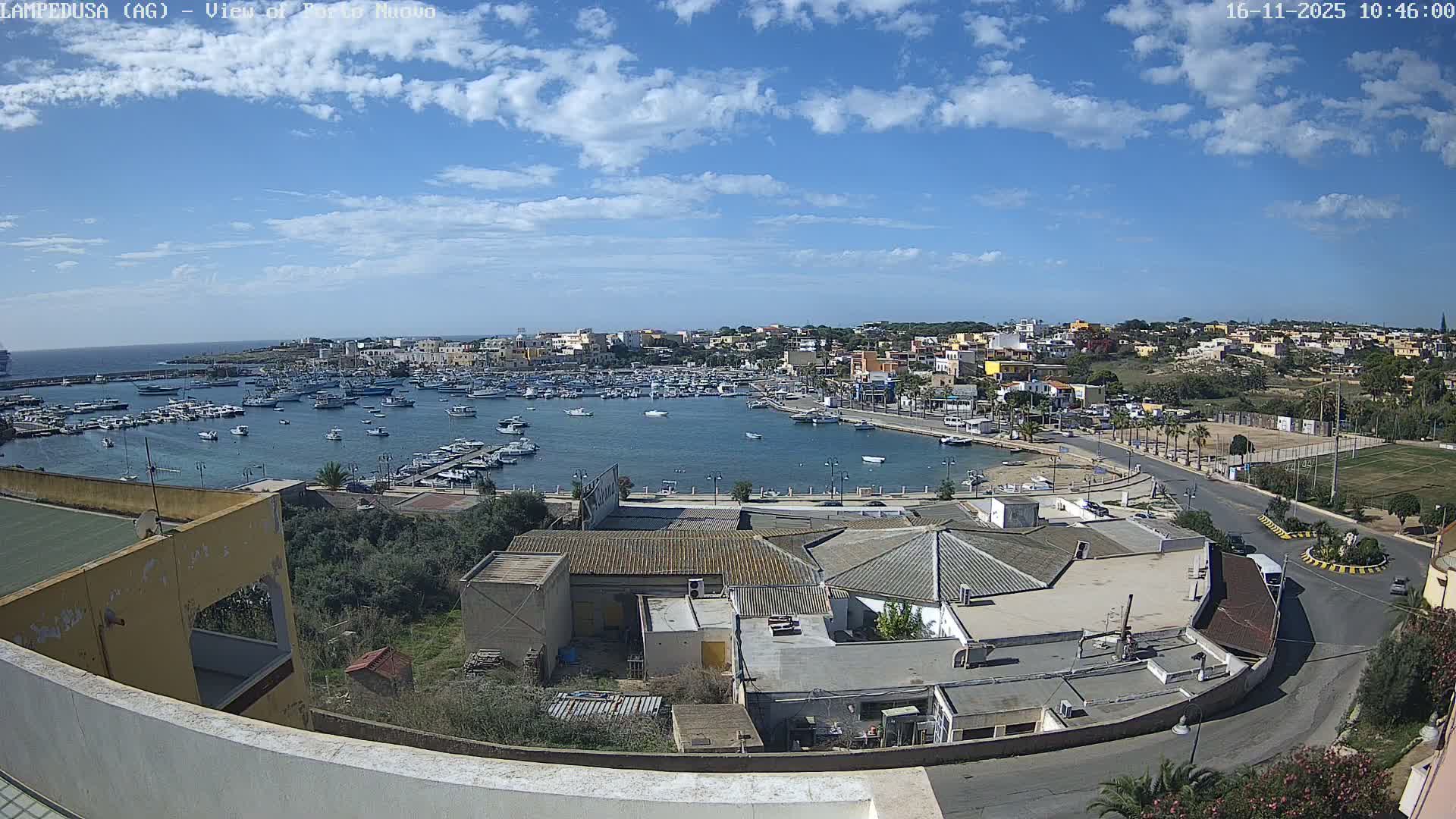 An elevated view shows a wide harbor densely packed with boats, flanked by a diverse coastal town and a winding waterfront road, all illuminated by bright sunshine under a blue sky with scattered white clouds.