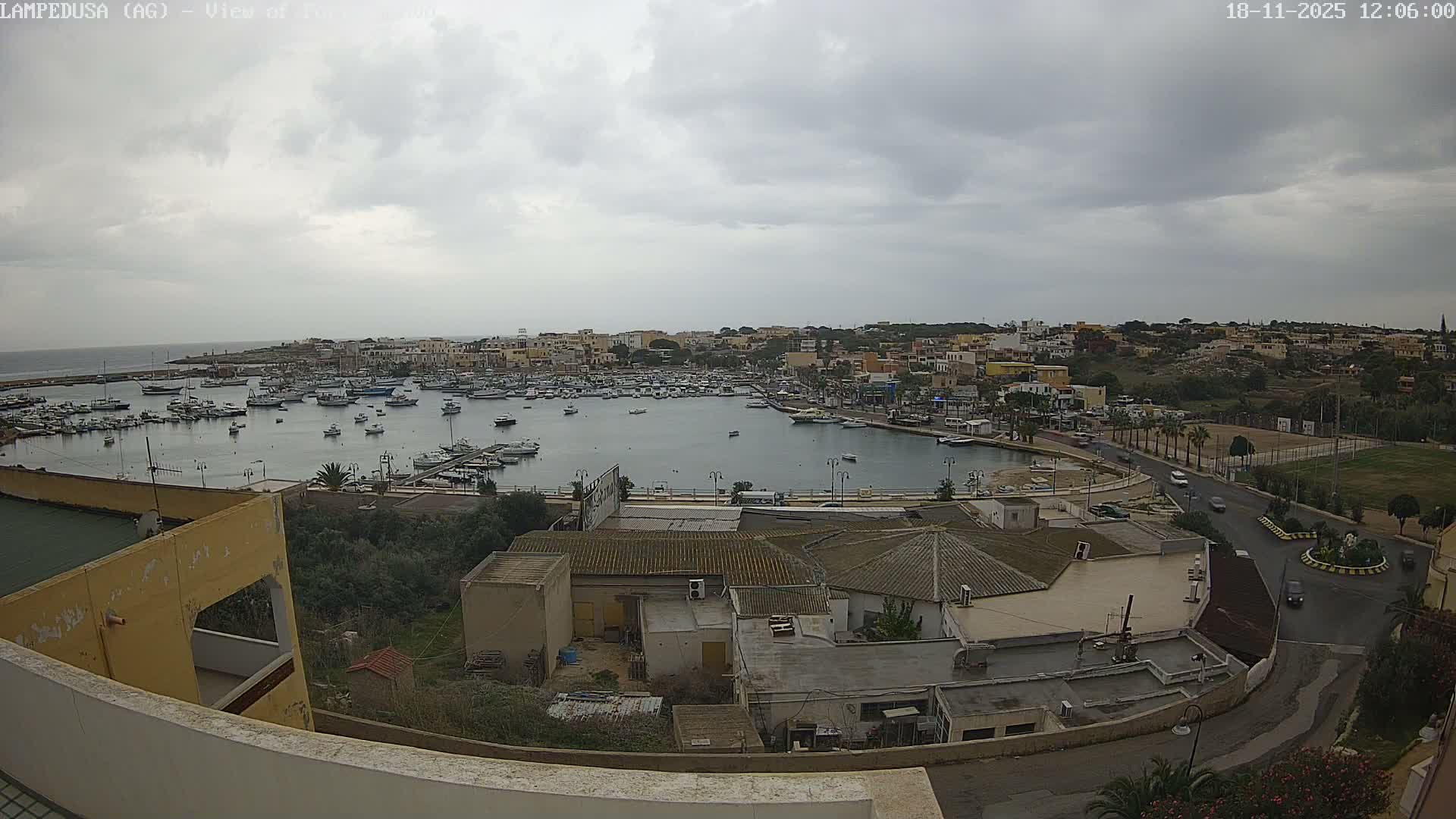 An elevated view shows a wide harbor densely packed with boats, flanked by a diverse coastal town and a winding waterfront road, all illuminated by bright sunshine under a blue sky with scattered white clouds.