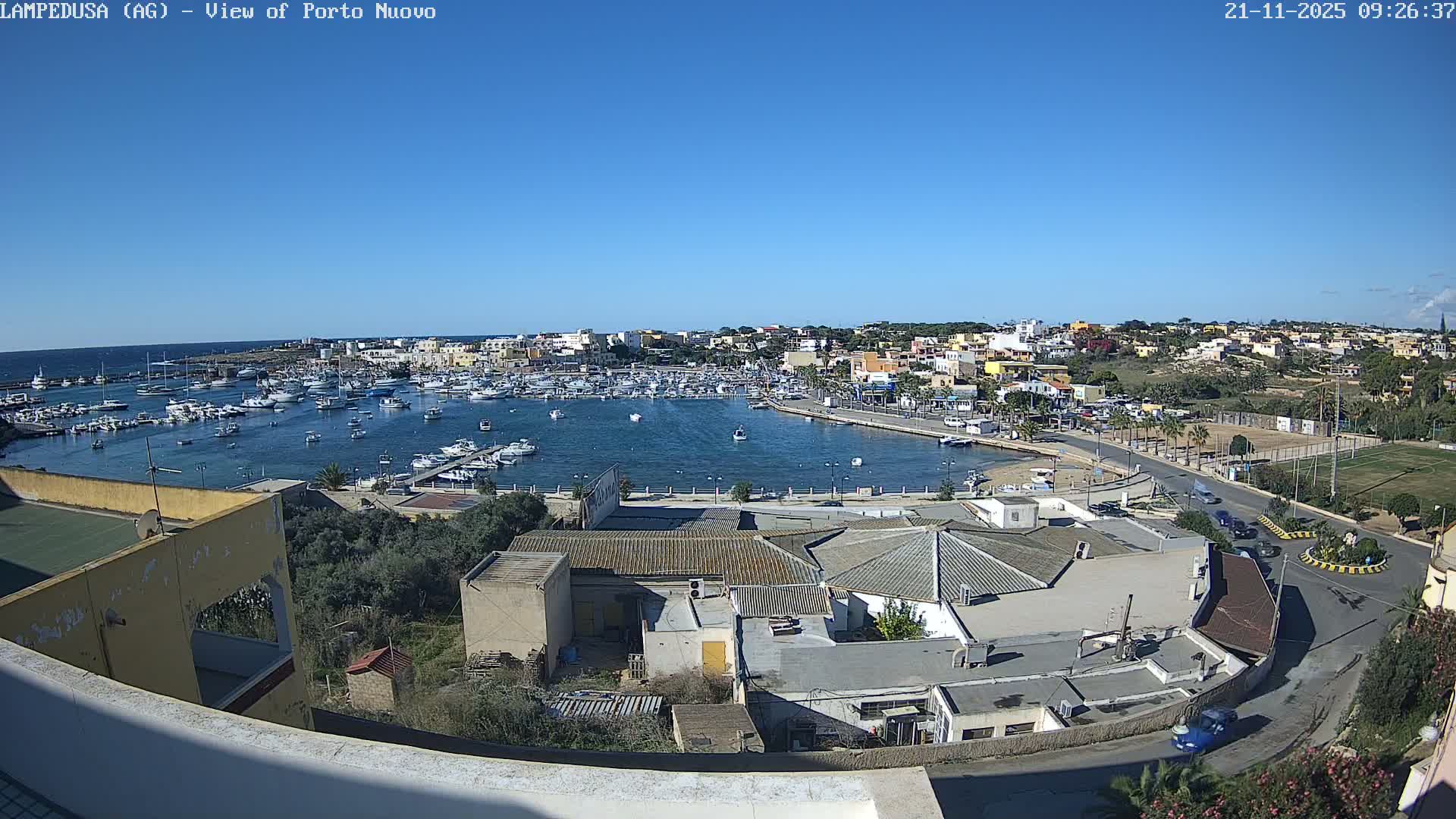 A clear, sunny day illuminates a busy harbor dotted with numerous boats, bordered by a vibrant coastal town with buildings, roads, and some green spaces.