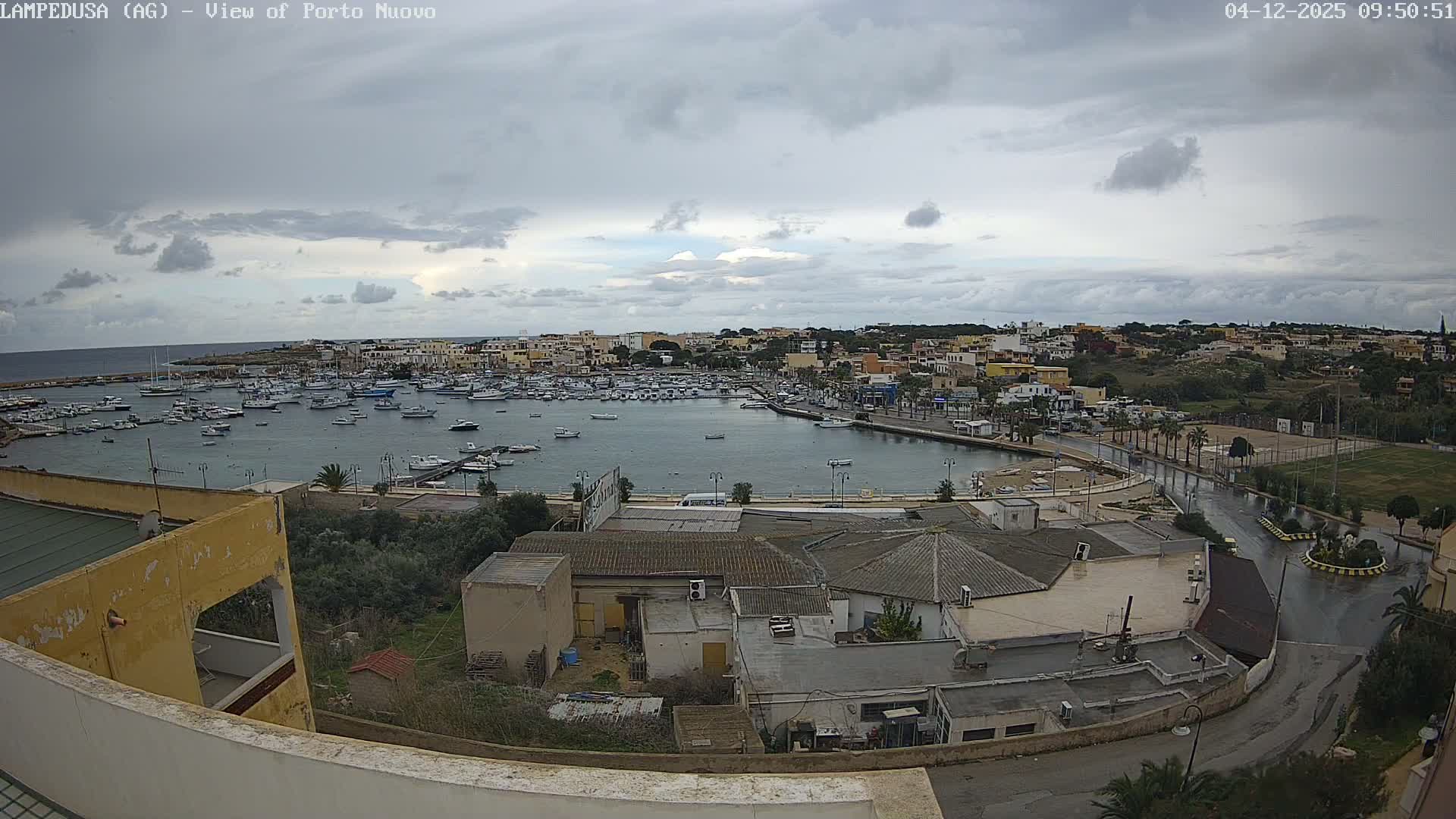 A panoramic view captures a bustling harbor filled with numerous boats, bordered by a colorful coastal town with a wet promenade, all under an overcast sky heavy with grey clouds.