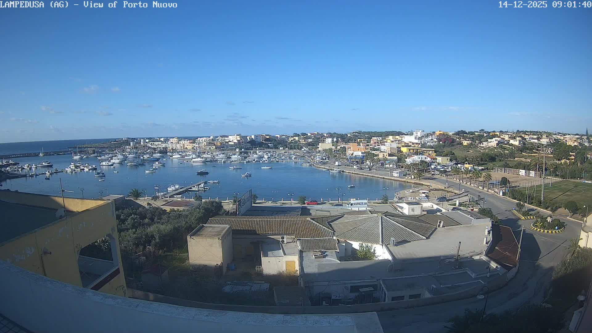 A sunny and clear day overlooks a bustling coastal town with a harbor filled with numerous boats, bordered by various buildings, roads, and green spaces, including a football field.