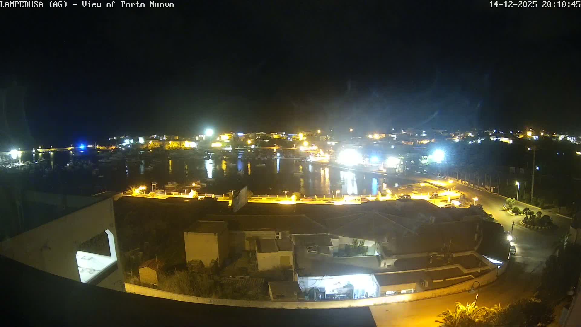 A sunny and clear day overlooks a bustling coastal town with a harbor filled with numerous boats, bordered by various buildings, roads, and green spaces, including a football field.