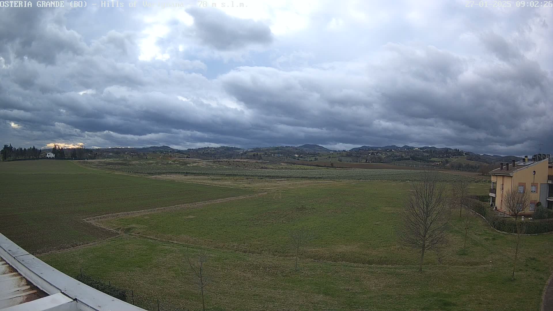 A sprawling, overcast landscape features flat green fields, distant rolling hills, and a few buildings, under a cloudy sky.