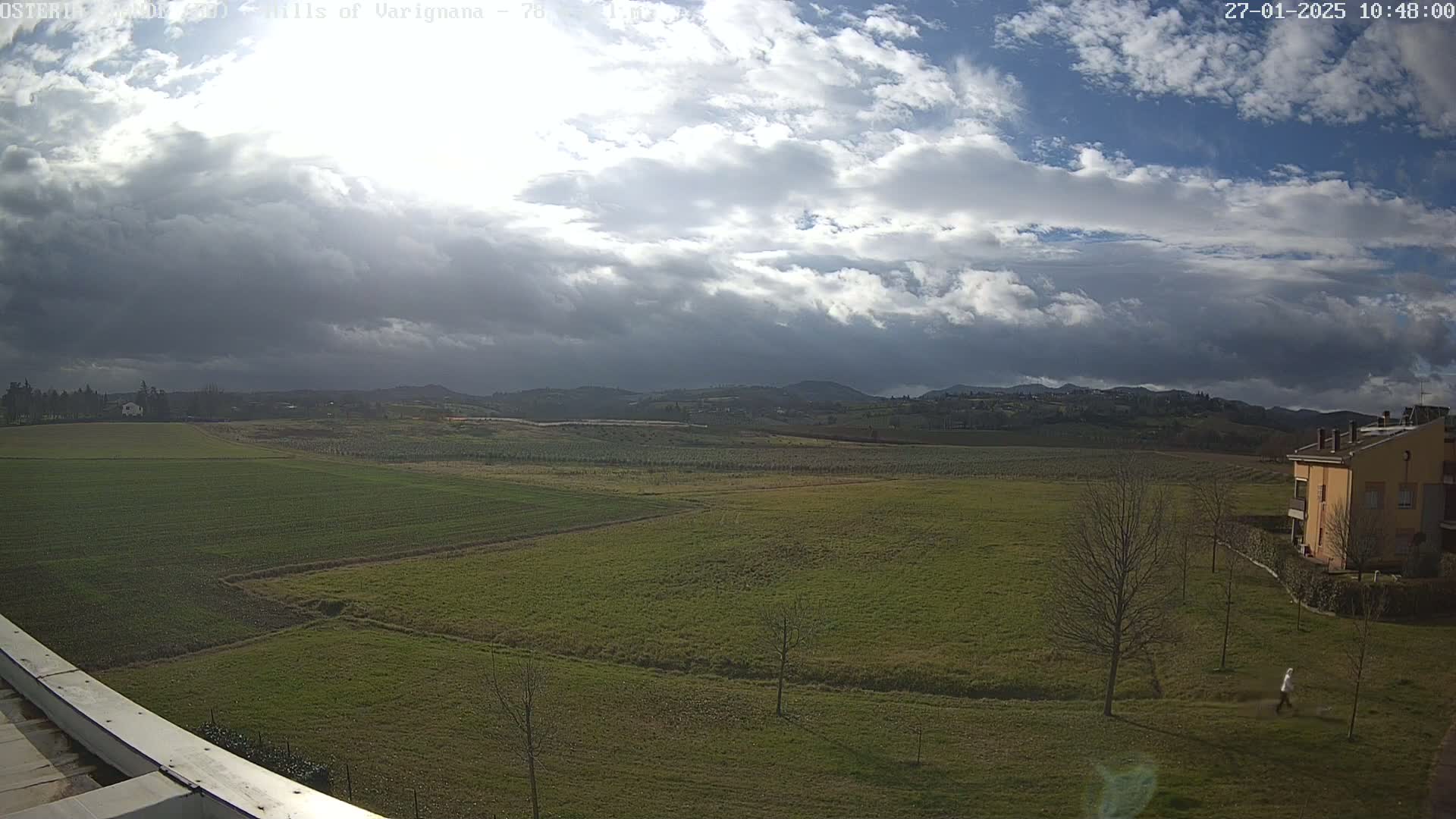 A partly cloudy day reveals a wide expanse of green fields and distant hills, with a few buildings visible near the bottom right.