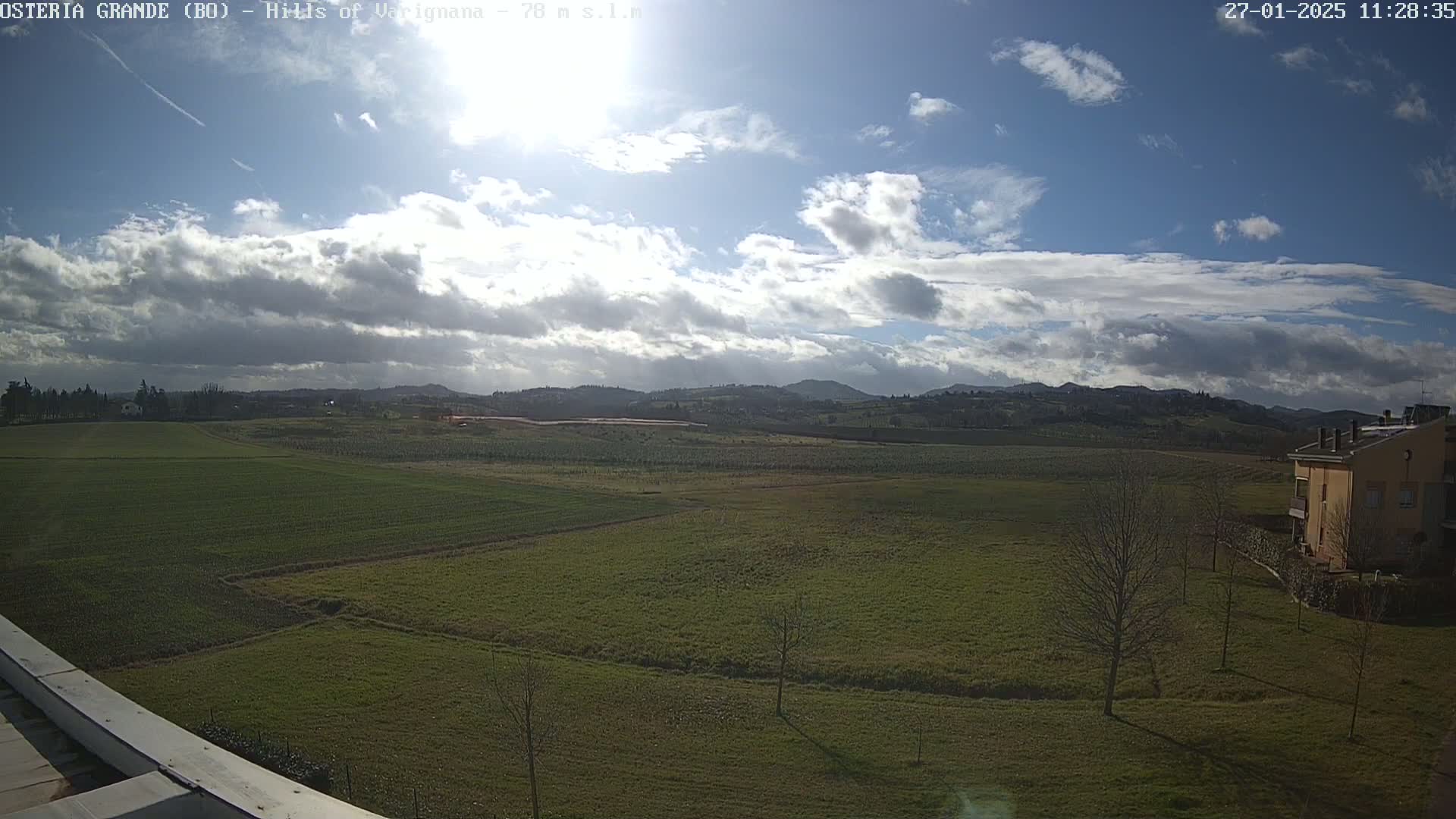 A partly cloudy day reveals a vast expanse of green farmland stretching towards rolling hills in the distance, with a building visible on the lower right.