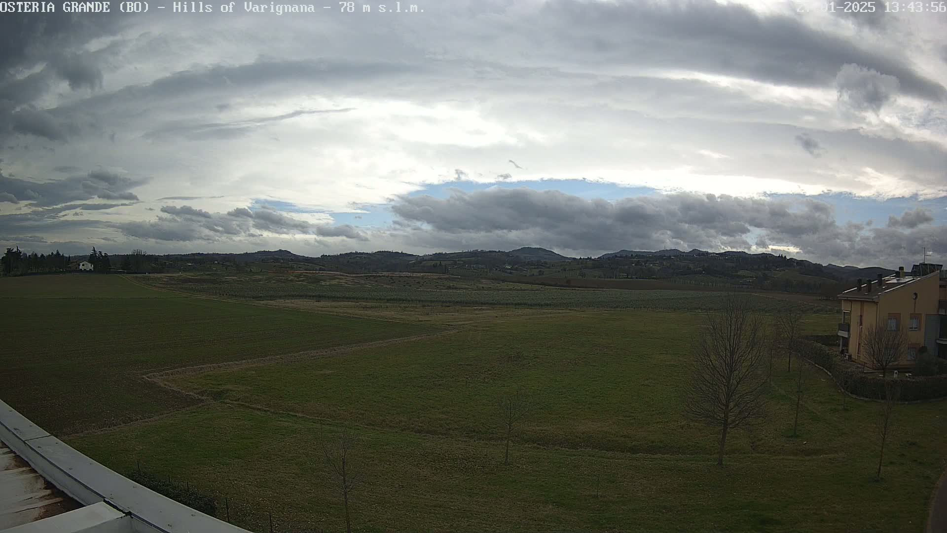 A mostly cloudy sky hangs over a flat expanse of green farmland, with distant hills and a few buildings visible.