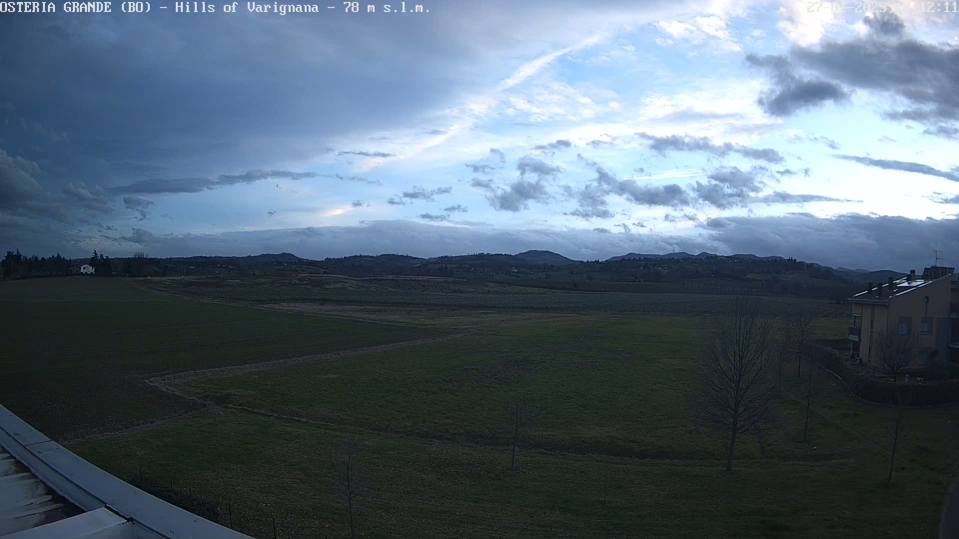 A wide, flat expanse of green fields stretches to a line of low hills under a mostly cloudy, but partly clear sky.