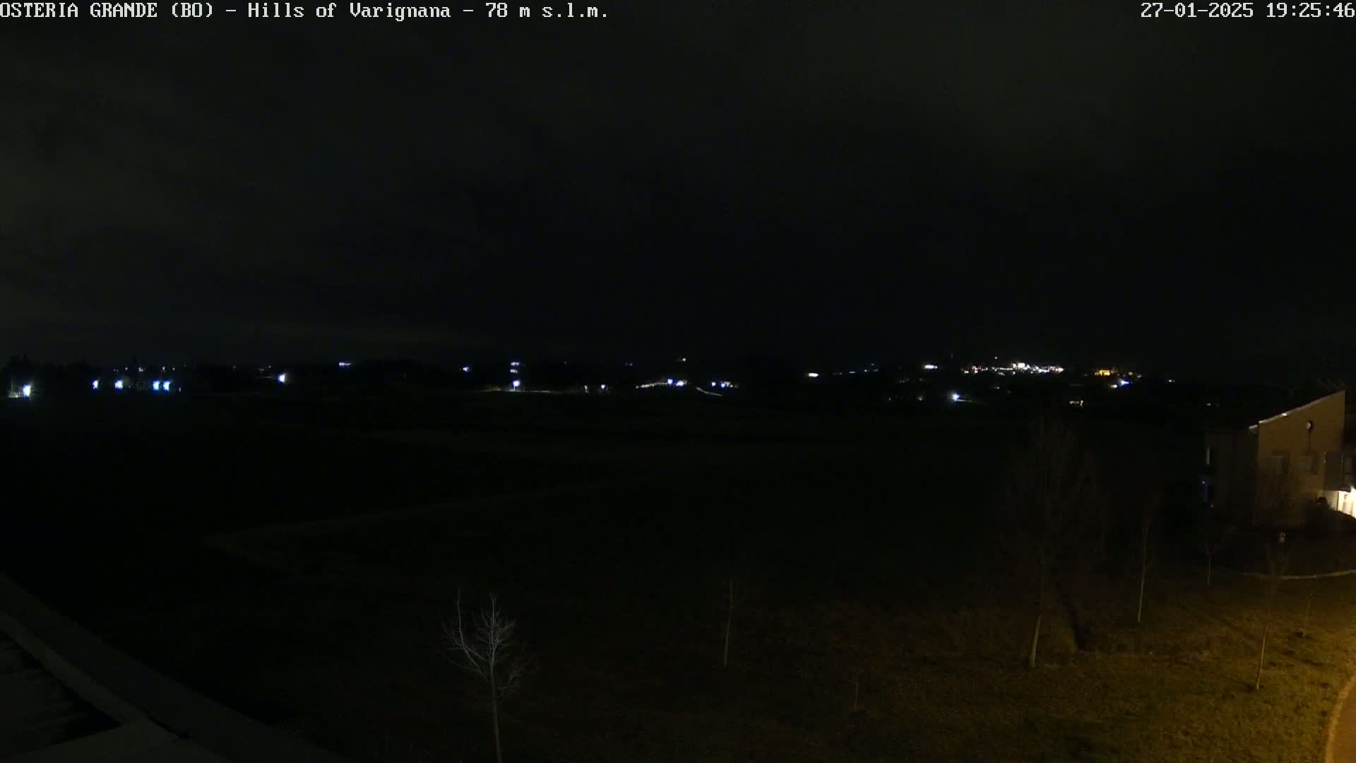 A dark, nighttime view of a sparsely lit landscape with a few distant lights and a building visible on the lower right.
