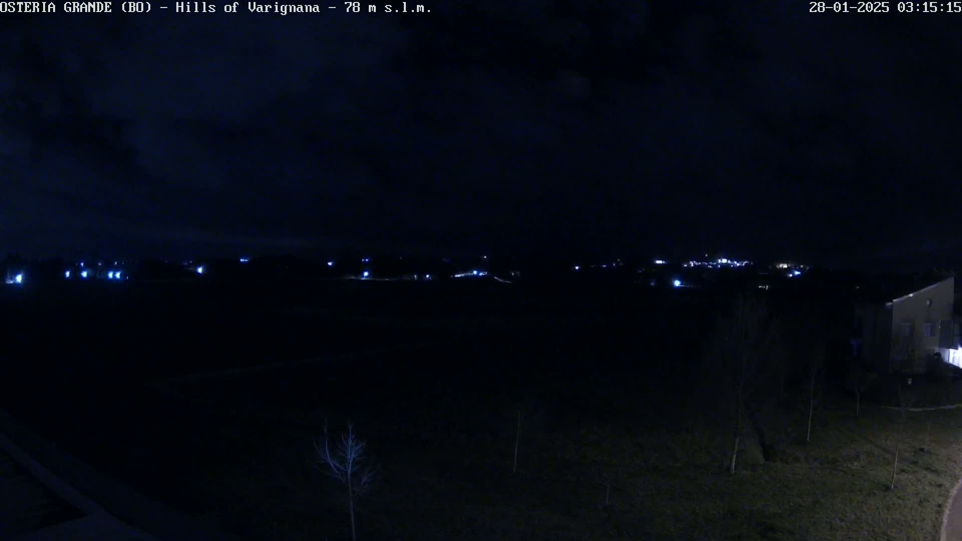 A nighttime view of a dark landscape with a few distant lights and a building partially visible on the right, under a mostly cloudy sky.