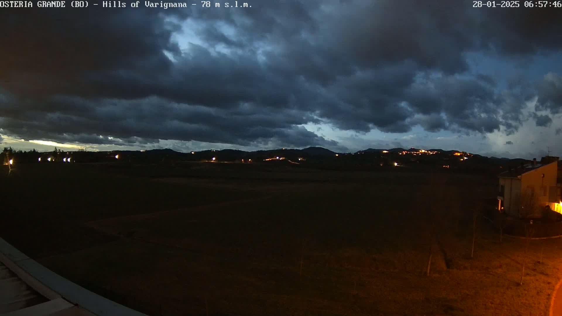 A dark, overcast night scene shows a flat, dark field extending to distant, dimly lit hills under a mostly cloudy sky.